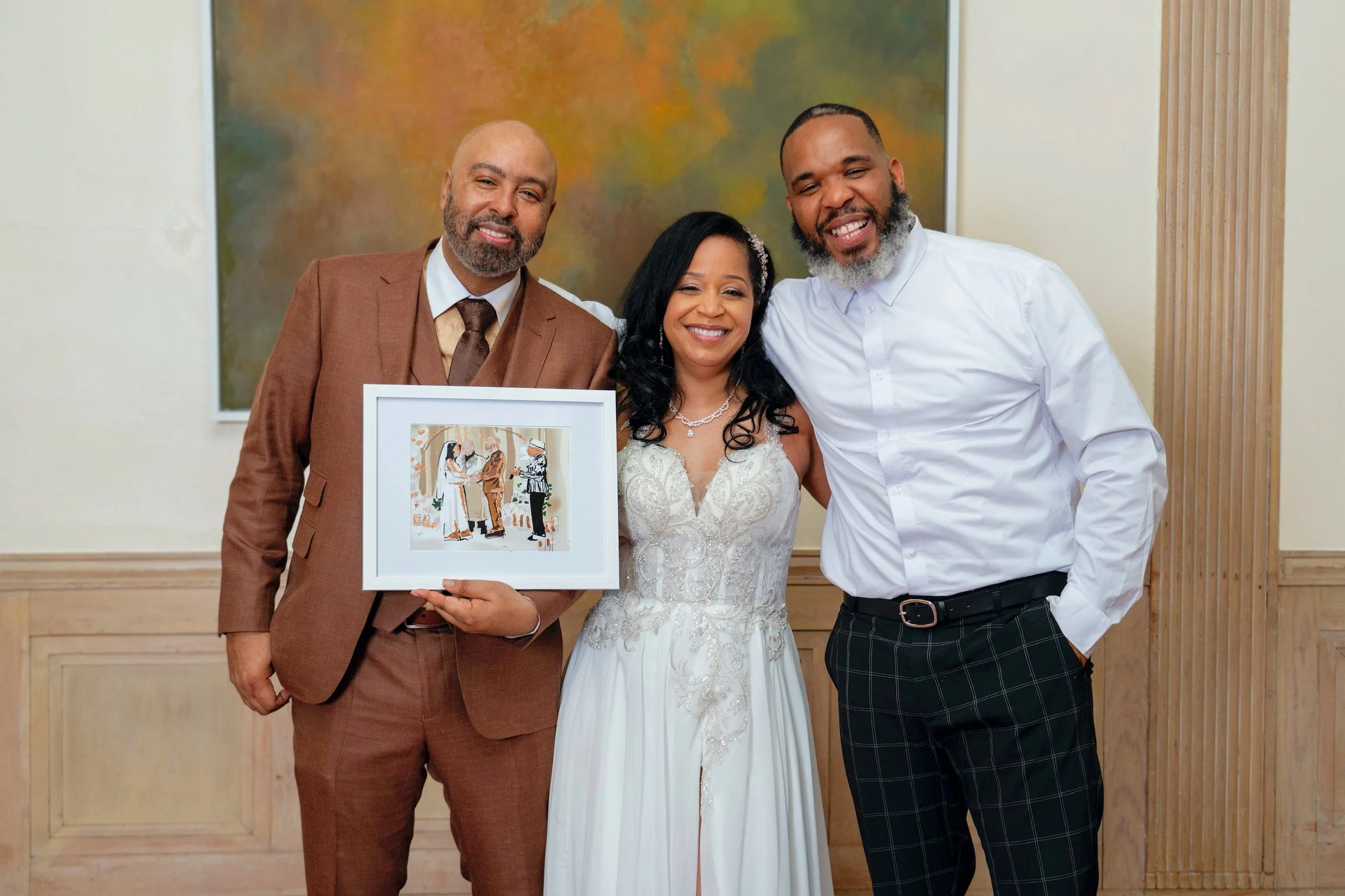 Three people smiling at a celebration, two men and a woman in a wedding dress, holding a framed artwork of an outdoor event with two women and a man, in front of a painted abstract background.