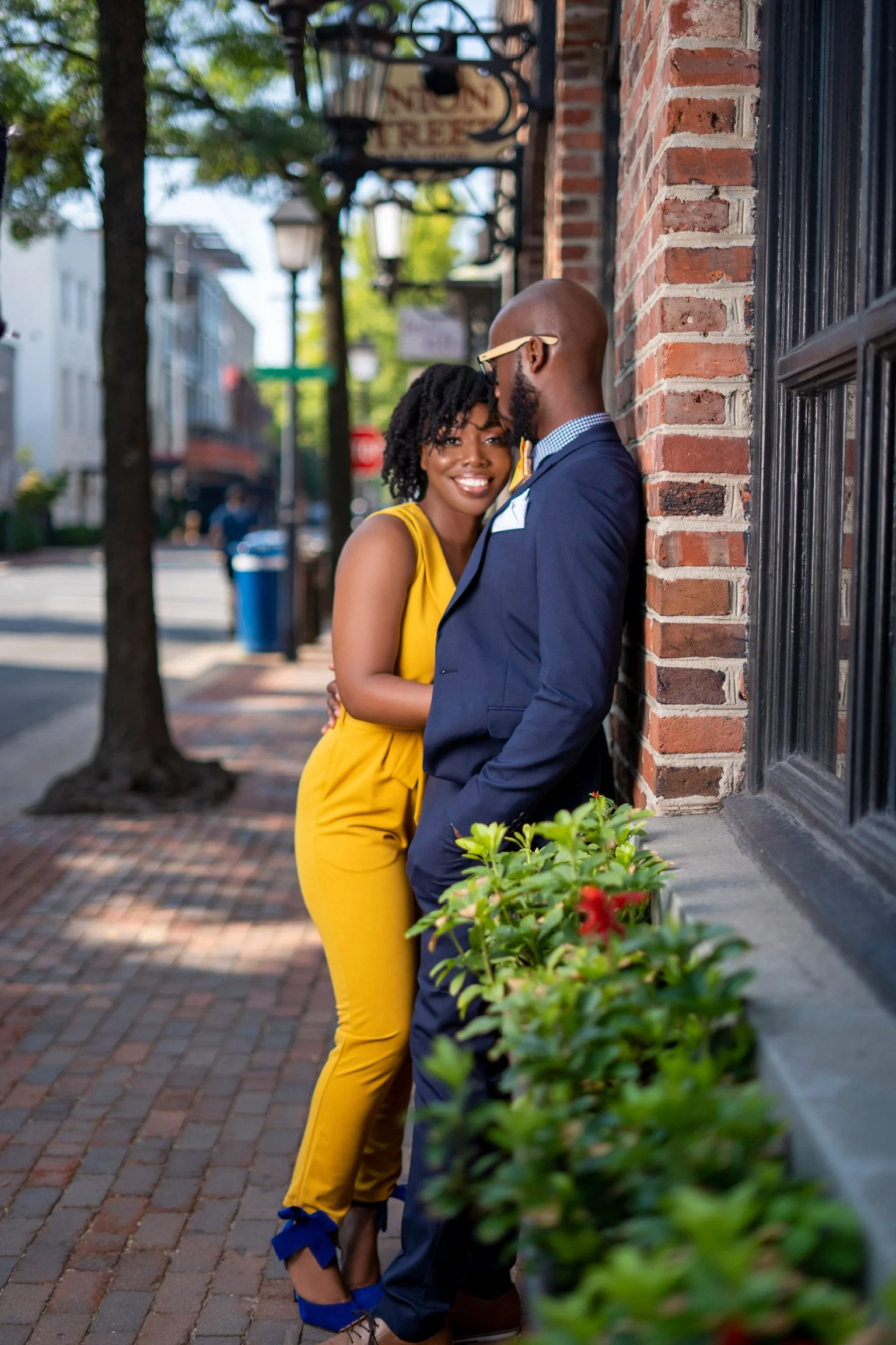 A happy woman in a yellow jumpsuit and blue high heels smiling and leaning close to a man in a navy blue suit, standing on a brick sidewalk in front of a brick wall with green plants in the foreground, in a city street setting.