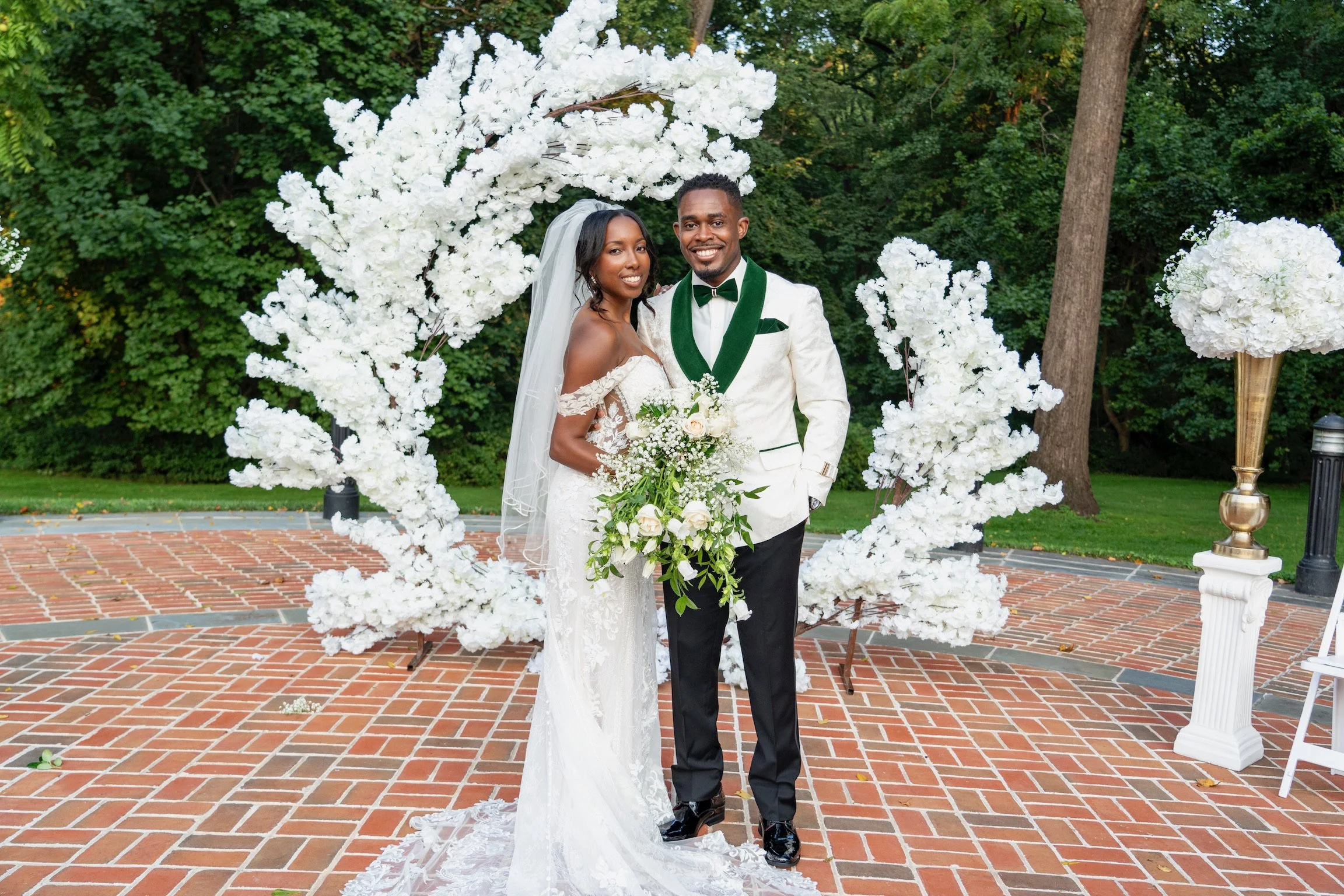 A bride and groom standing together outdoors in front of a white floral arch, surrounded by greenery, on a brick-paved area during their wedding celebration.