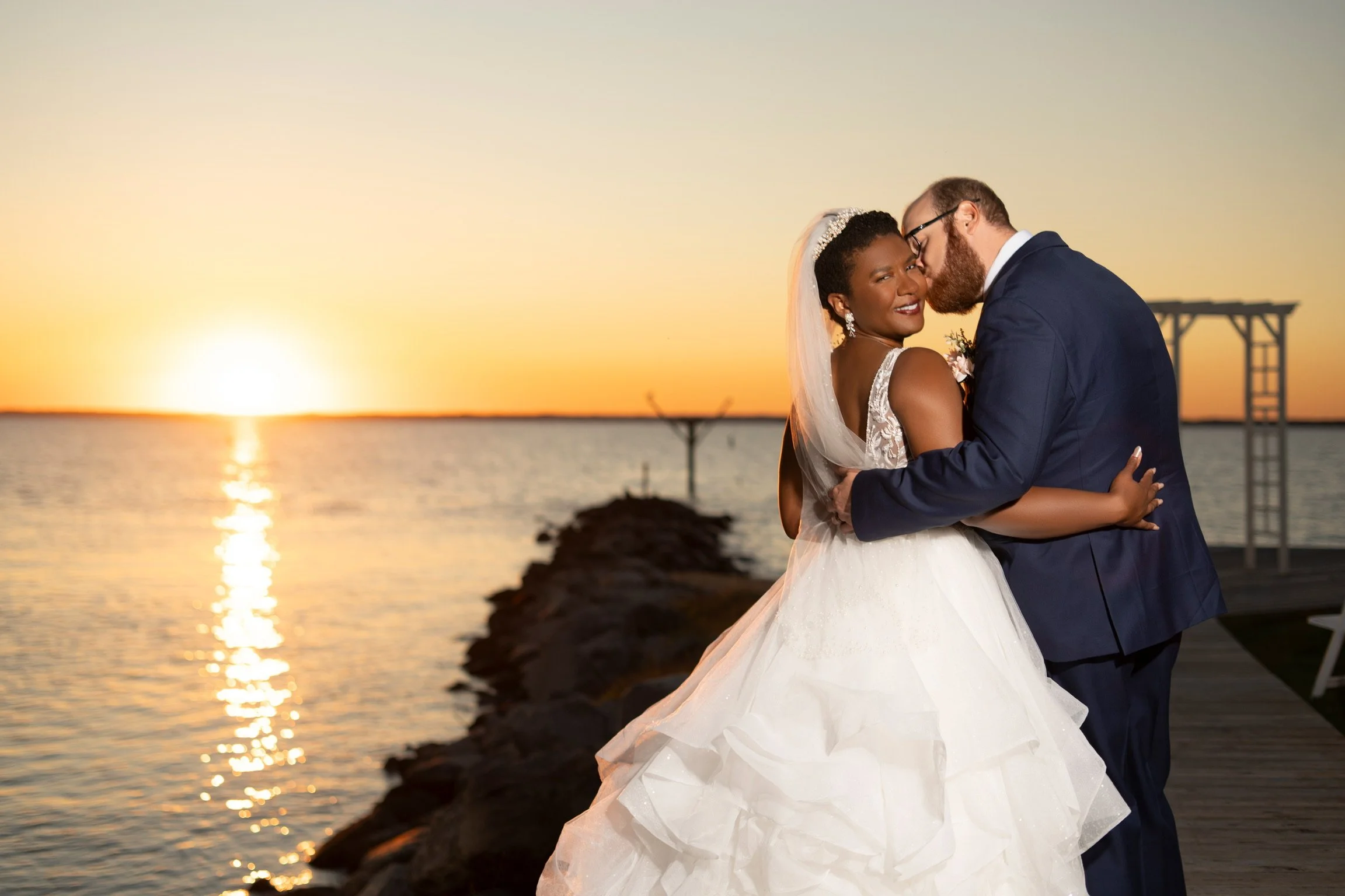A newlywed couple is embracing on a dock at sunset by the water. The bride is wearing a white wedding gown and veil, and the groom is in a navy suit. The sky is orange and yellow, and the sun is setting on the horizon.