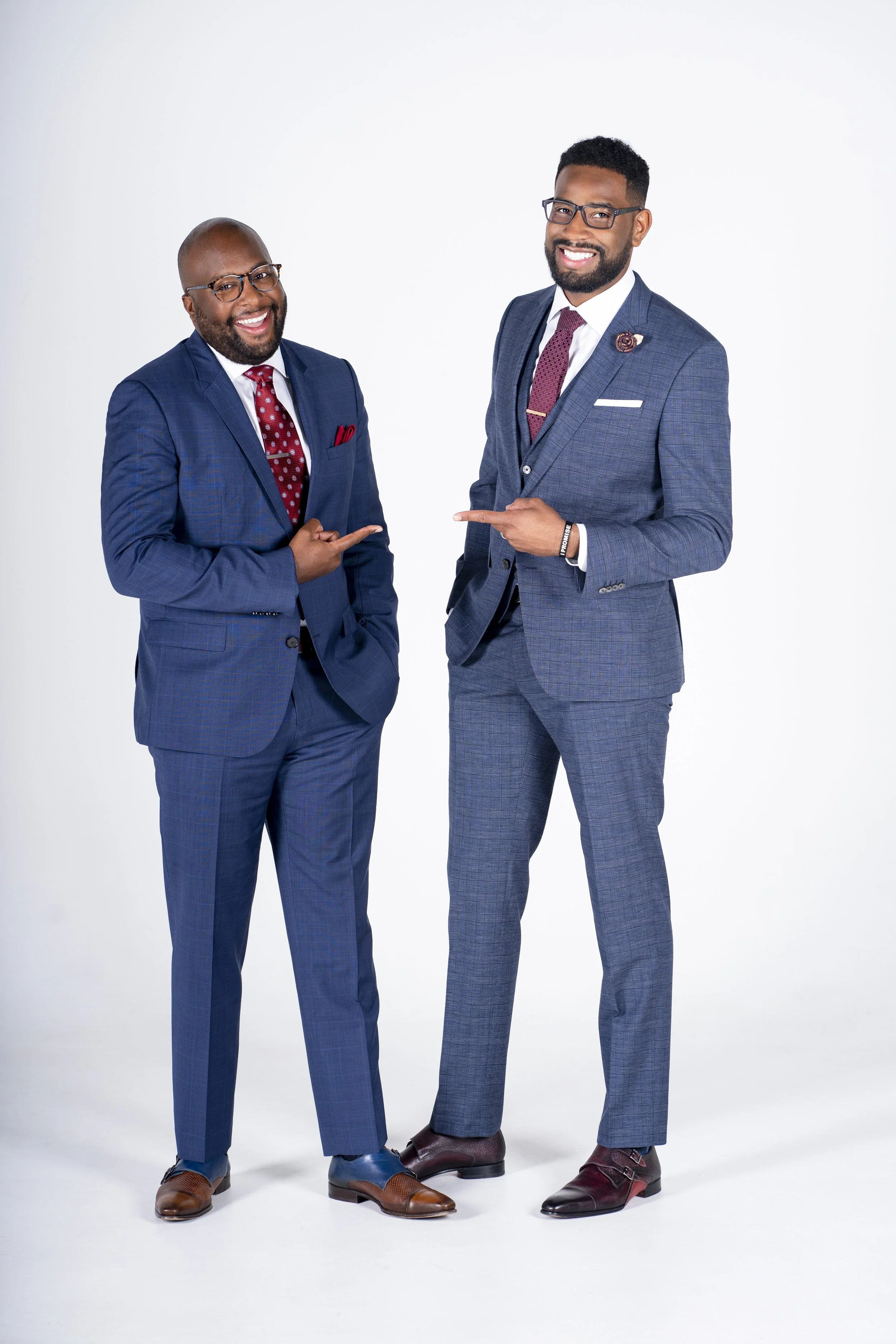 Two men in suits smiling and pointing at each other, standing against a white background.