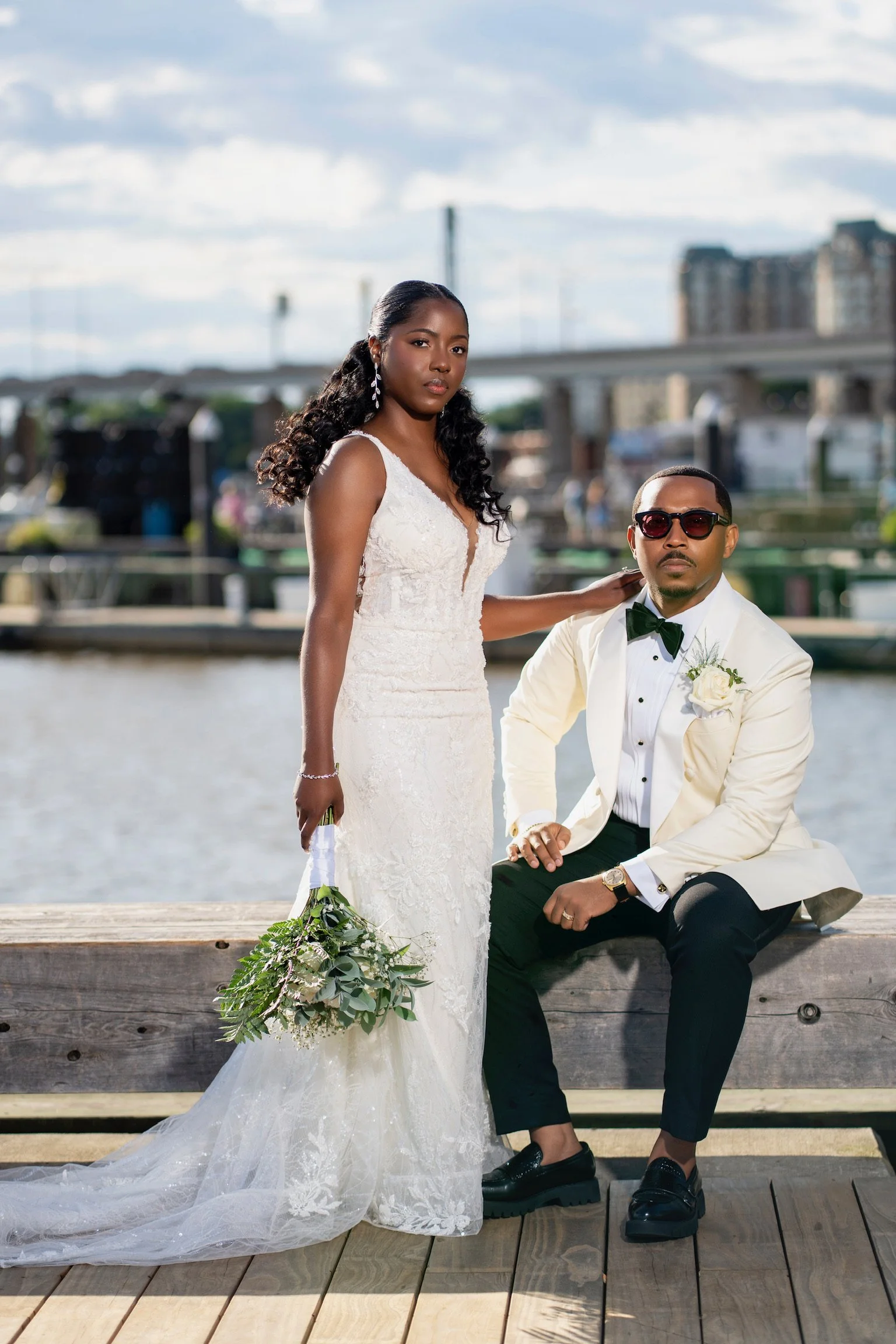 A bride wearing a white lace wedding gown and holding a bouquet stands beside a groom in a white tuxedo jacket, black pants, sunglasses, and a bow tie, sitting on a dock near the water with a cityscape and bridge in the background.