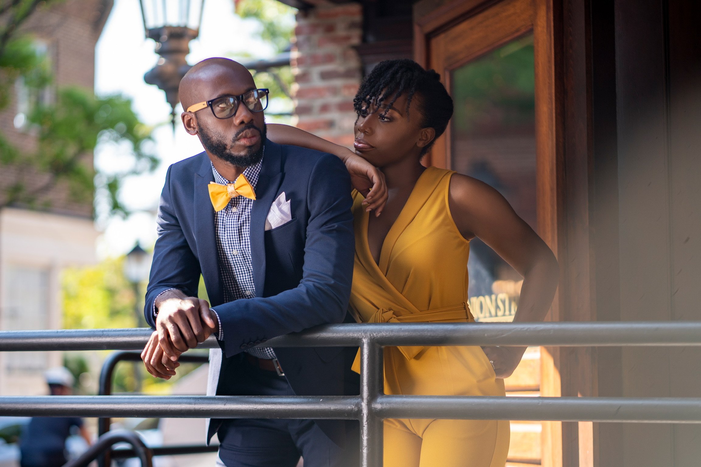 A man in a blue suit with glasses, a checkered shirt, and a yellow bow tie is leaning on a railing, looking away. A woman in a yellow dress leans her head on his shoulder, looking at him. They are outside, next to a brick building with trees in the b