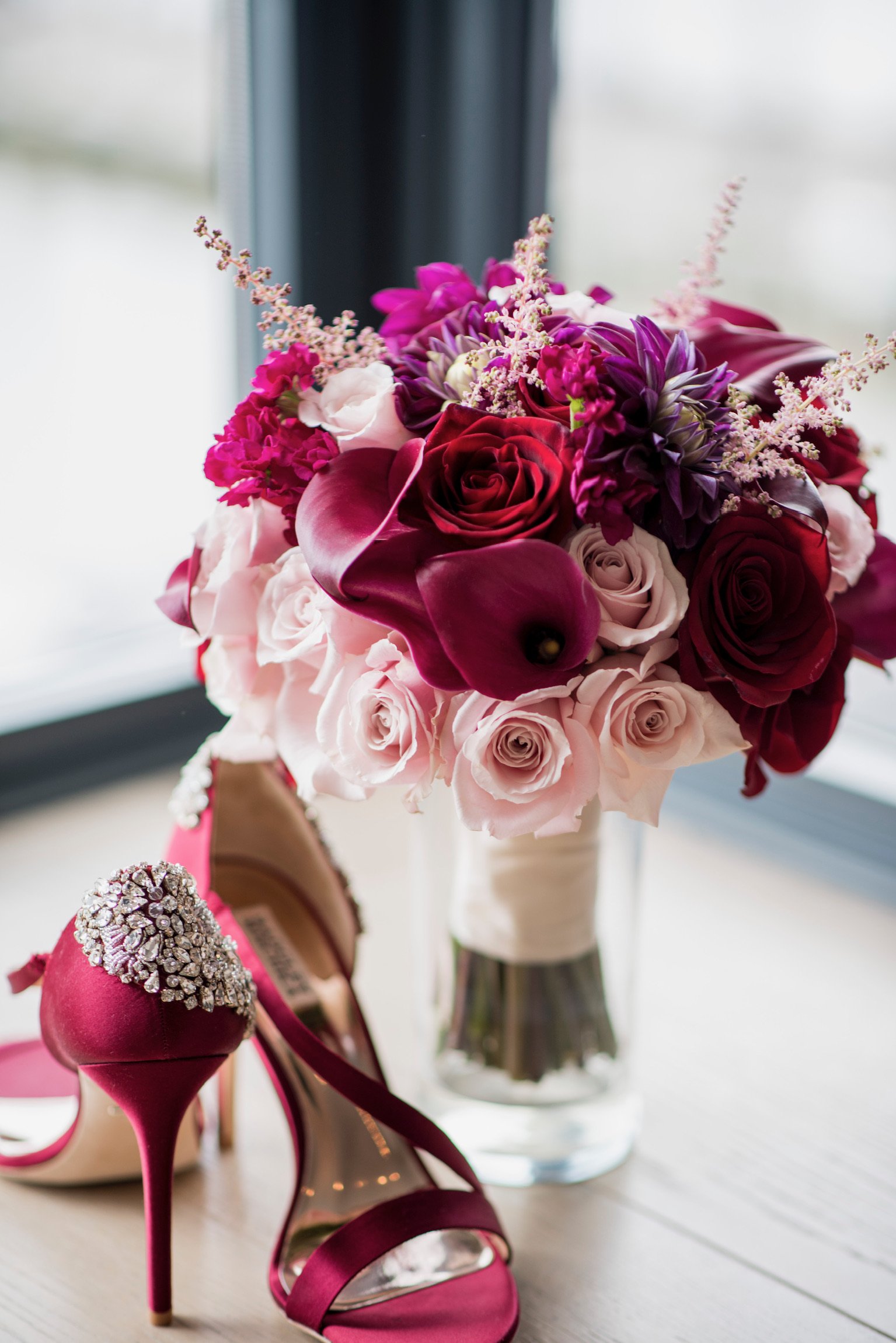 A bouquet of pink, white, and purple flowers in a glass vase, with pink high heels with rhinestone details beside it, on a wooden surface near a window.