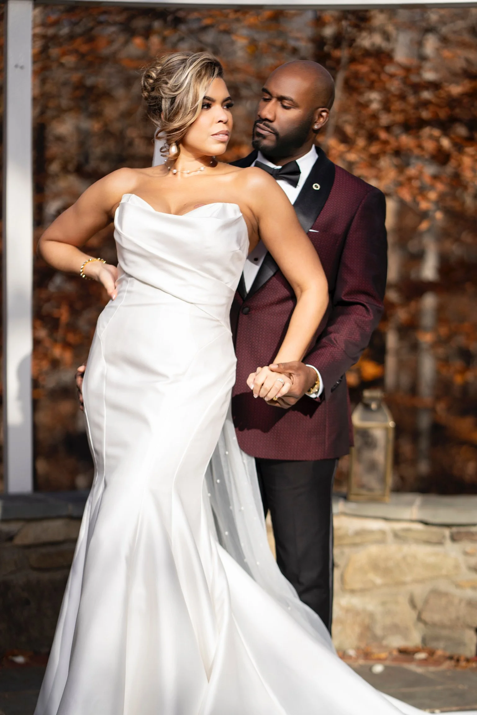 A bride and groom dancing on their wedding day, with the bride in a strapless white gown and the groom in a burgundy tuxedo jacket and black pants, in an outdoor setting with autumn leaves in the background.