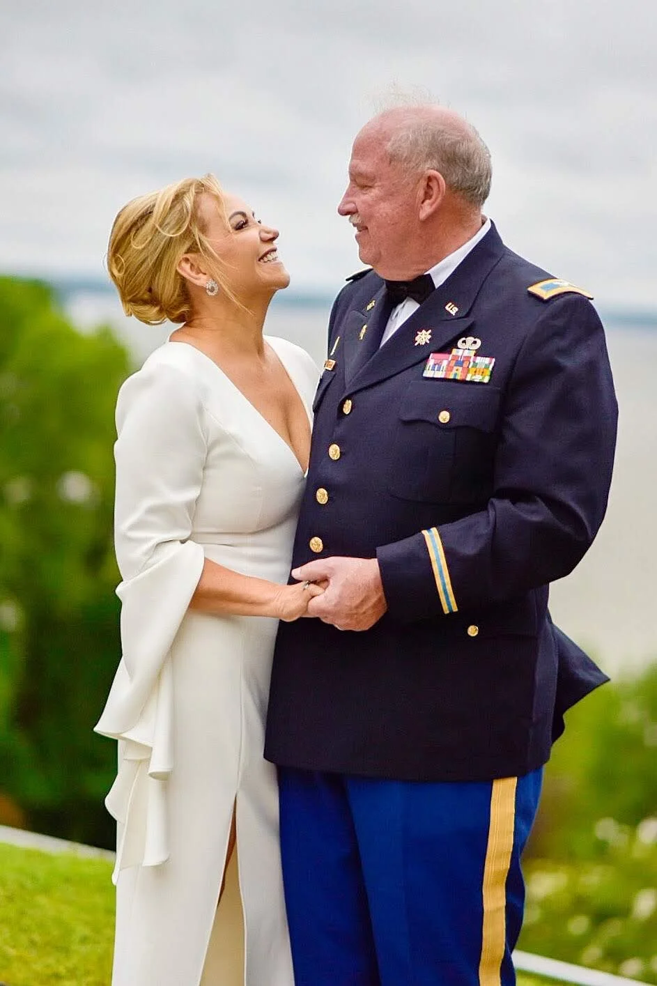 A woman in a white dress and a man in a U.S. military uniform are smiling and holding hands, standing outdoors with a blurred natural background.