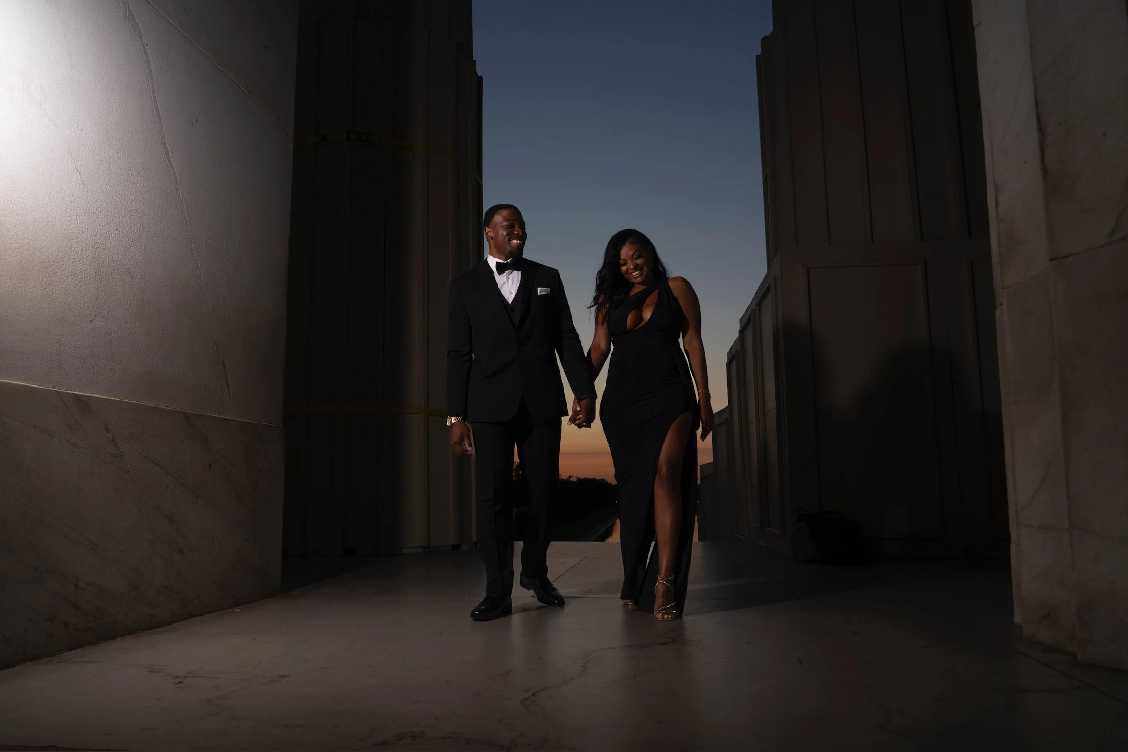 A couple dressed in formal evening attire walking outdoors during sunset, holding hands and smiling.