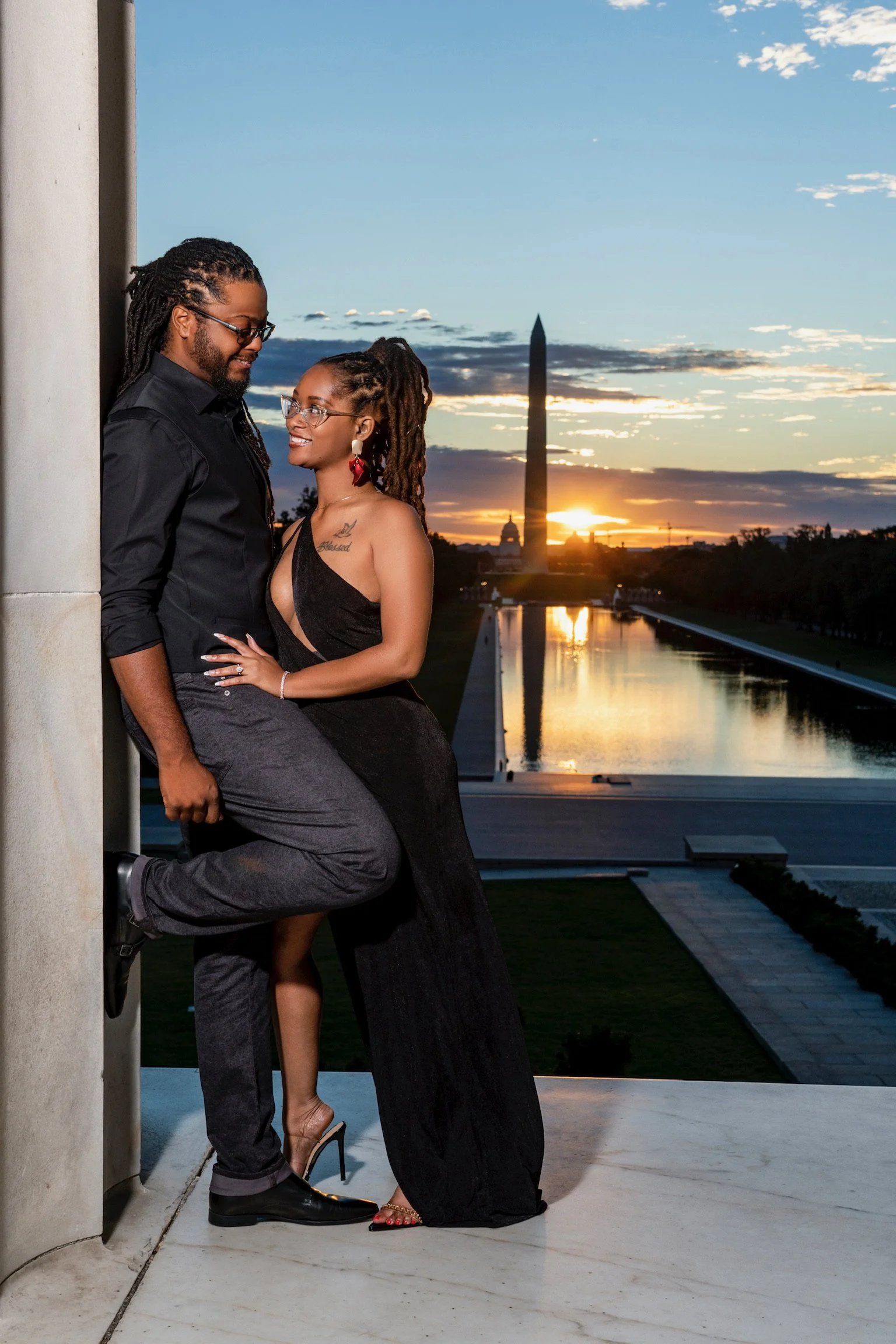 A couple dressed in formal attire poses together on a balcony with the Washington Monument and the reflecting pool at sunset in the background.