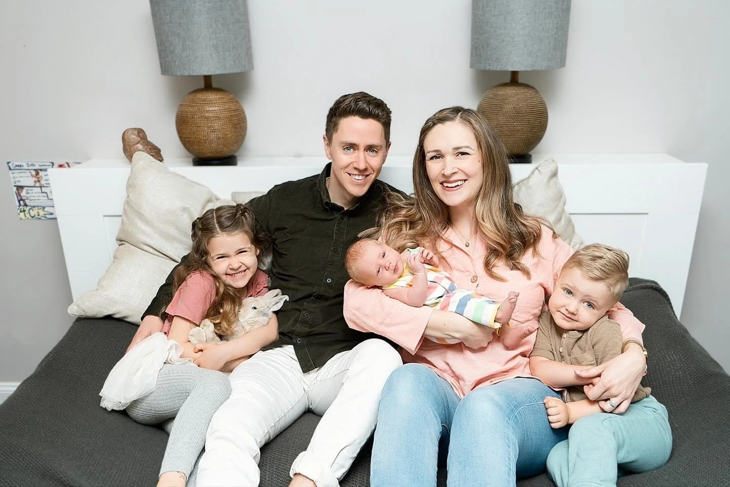 A happy family of five sitting on a couch, including a mother, father, and three young children, smiling and cuddling.