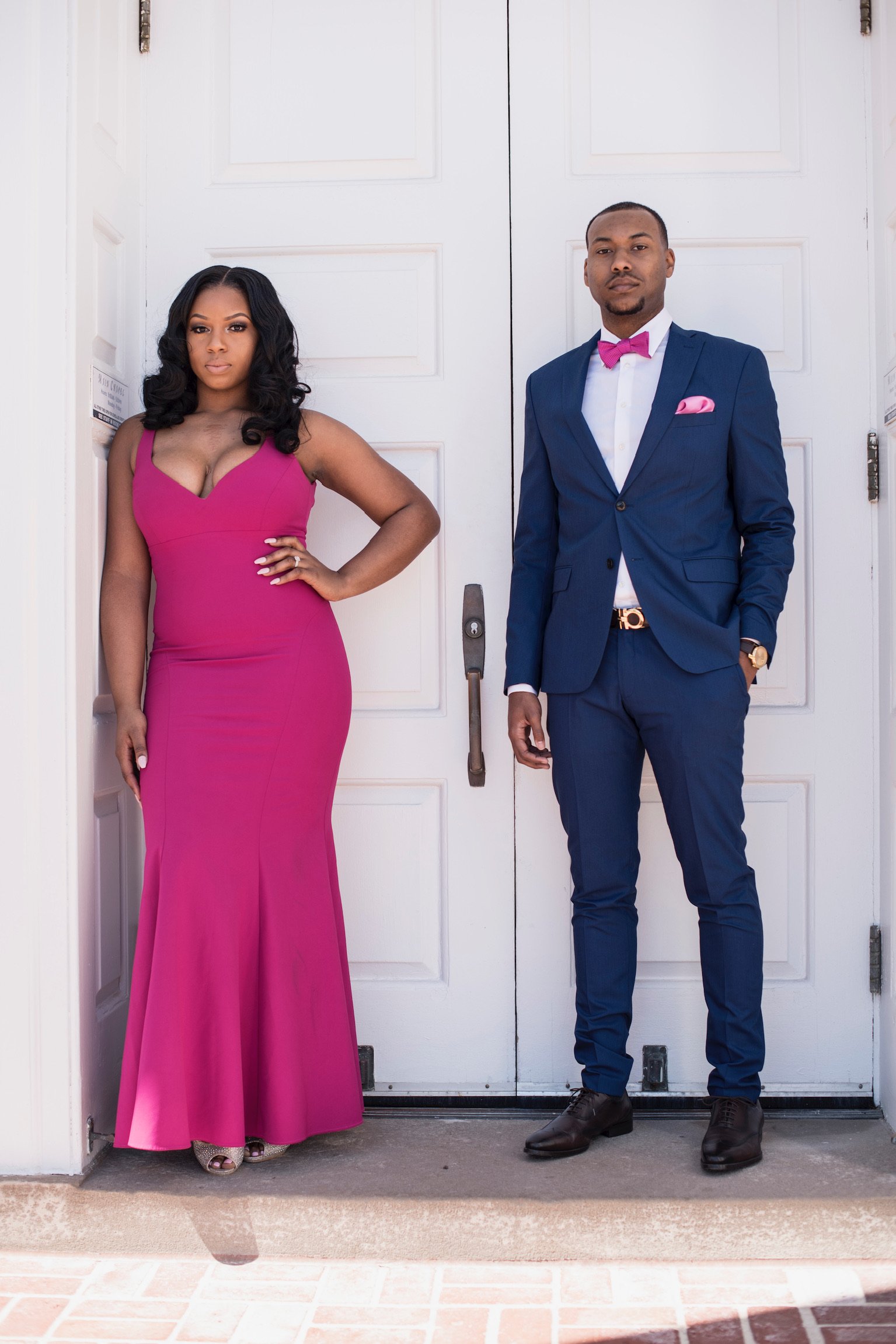 A woman in a pink gown and a man in a blue suit with a pink bow tie stand side by side in front of white doors.