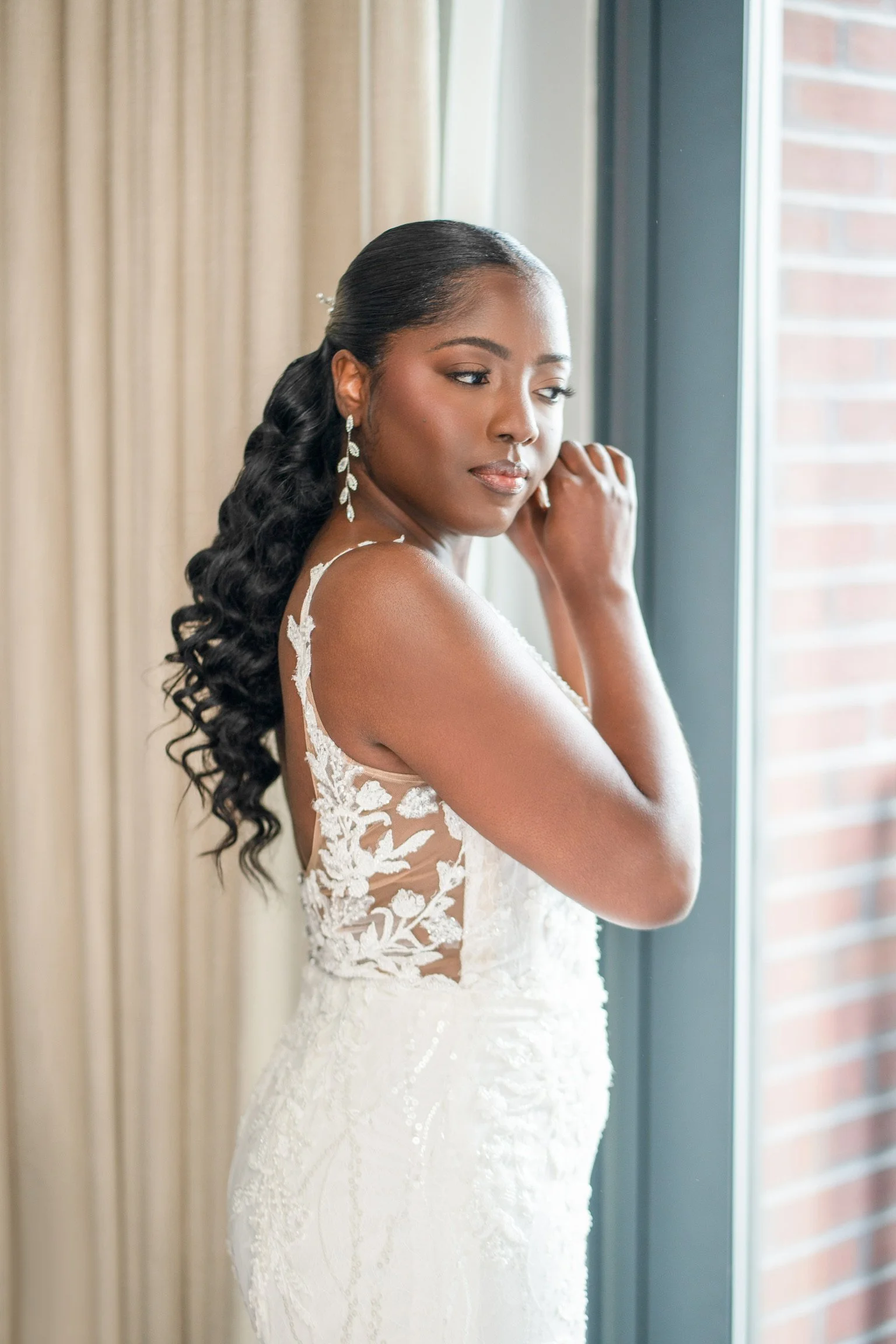 A bride in a white lace wedding dress standing by a window, touching her face, with a calm expression.