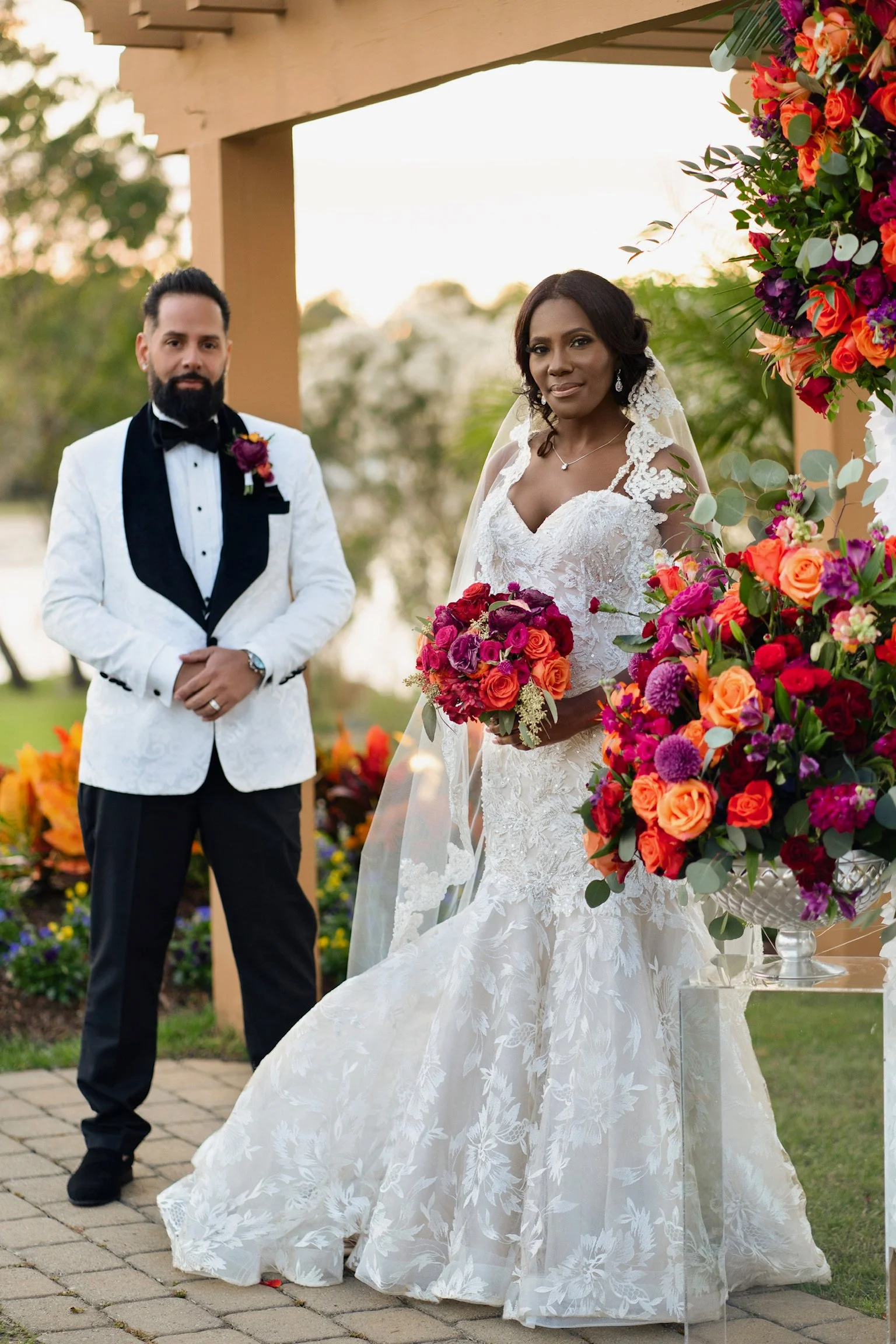 A bride and groom standing outdoors during his wedding ceremony, with colorful flowers decorating the surroundings.