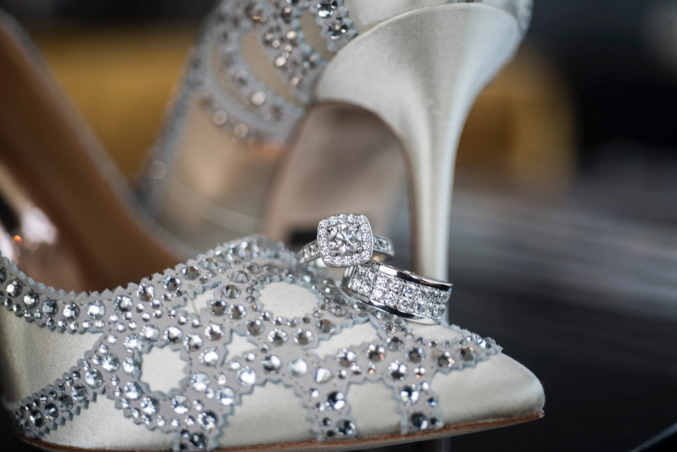 A diamond engagement ring and wedding band placed on a white satin shoe with reinforced beaded and rhinestone embellishments, with a white satin high-heeled shoe in the background.