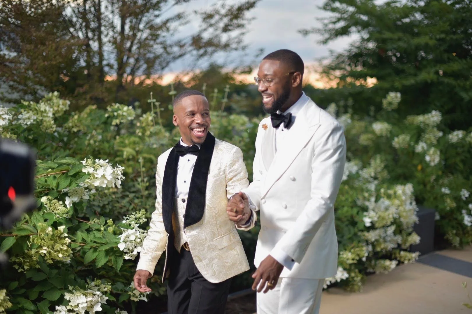 Two men dressed in tuxedos, one in white and one in gold, smiling and holding hands outdoors with greenery and white flowers in the background during the evening.