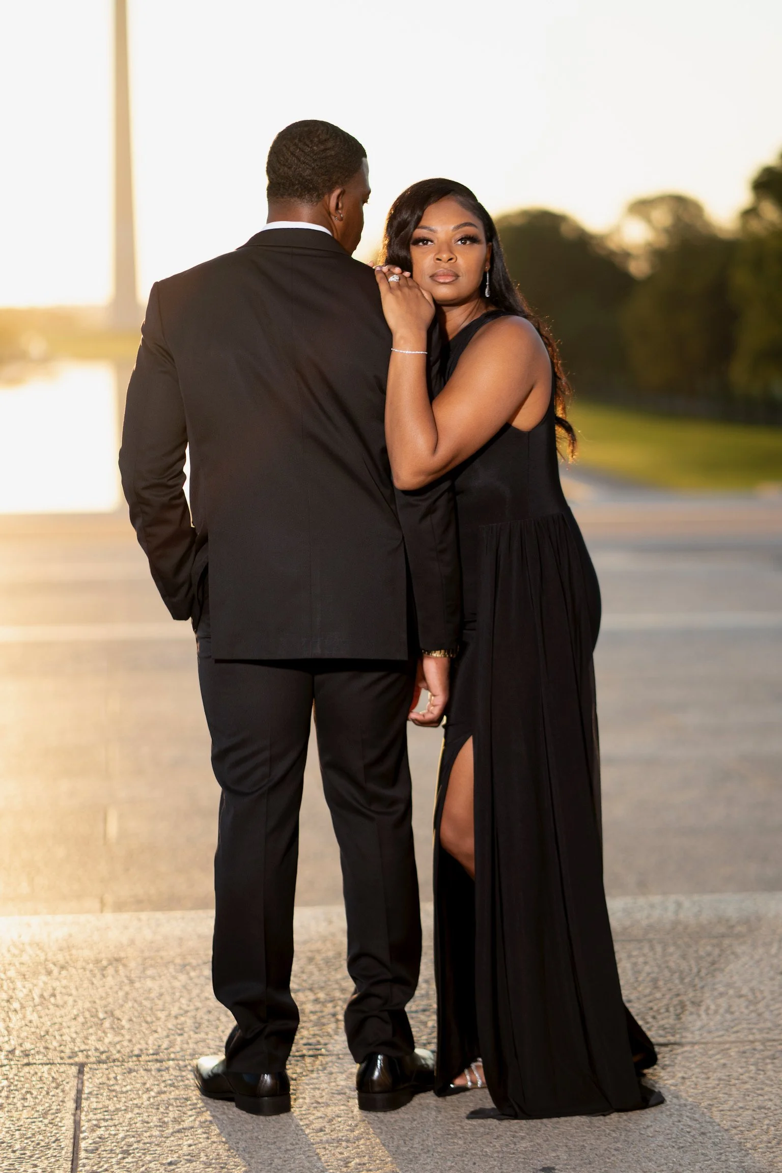 A couple dressed in formal black attire, standing closely outdoors during sunset, with the woman gently touching the man's shoulder and looking at the camera.
