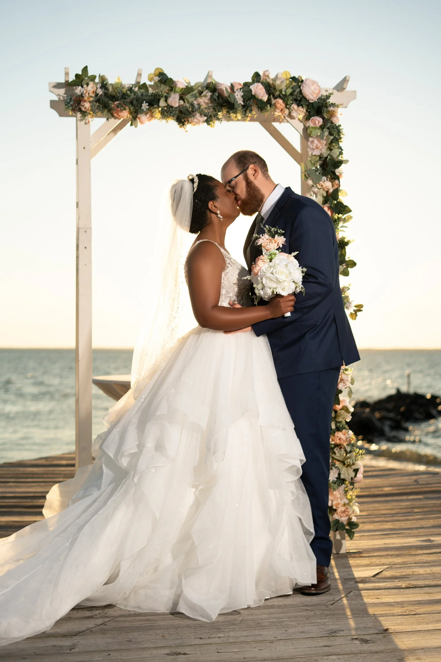 A bride and groom kissing during their beach wedding, standing on a wooden pier with a floral arch behind them and the ocean in the background.