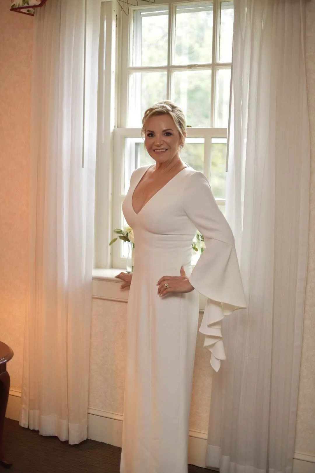 A woman in a white dress with long sleeves and ruffled cuffs, standing in front of a window with white curtains, smiling and posing with her hands on her hips.