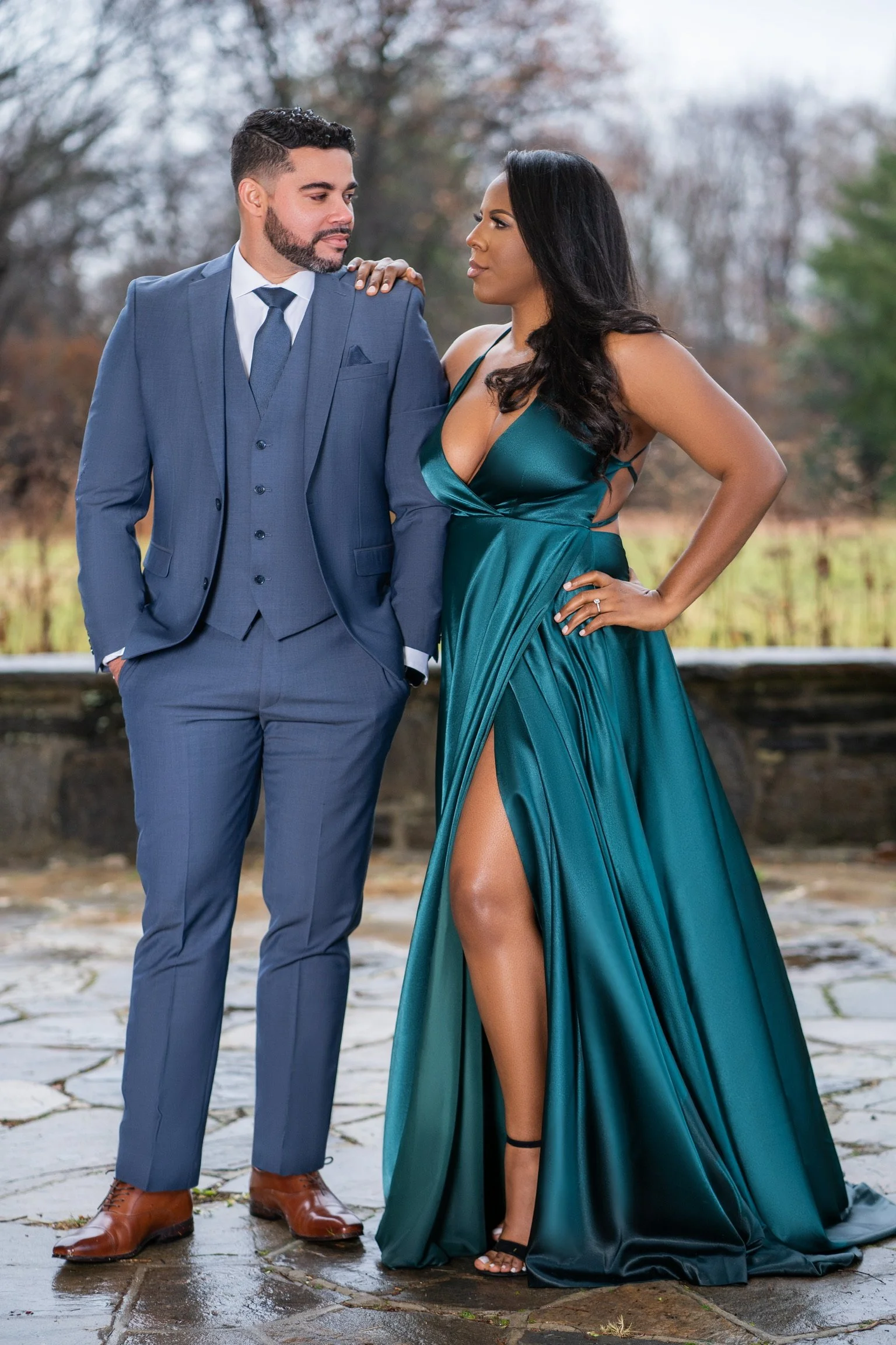 A man and woman standing outdoors on a stone path. The man is wearing a blue suit, white shirt, and brown shoes, with his hands in his pockets. The woman is wearing a teal satin dress with a slit, black heels, and has one hand on her hip. They are lo