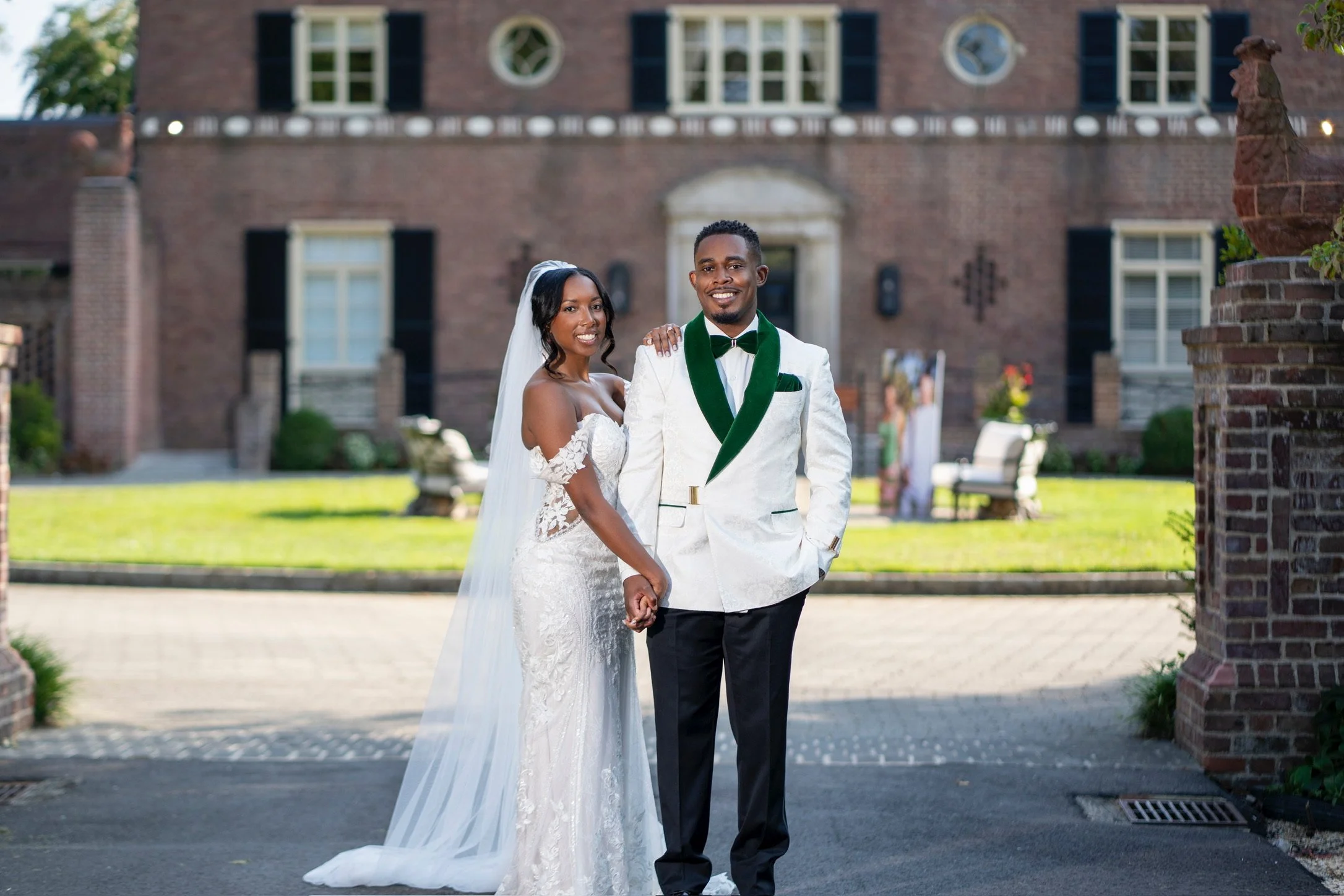 A newlywed couple holding hands and smiling outdoors in front of a brick building. The bride is wearing a white wedding gown with lace details and a veil, while the groom is dressed in a white jacket with black pants and a green and black bow tie. The background features a grassy area and a brick building with windows.