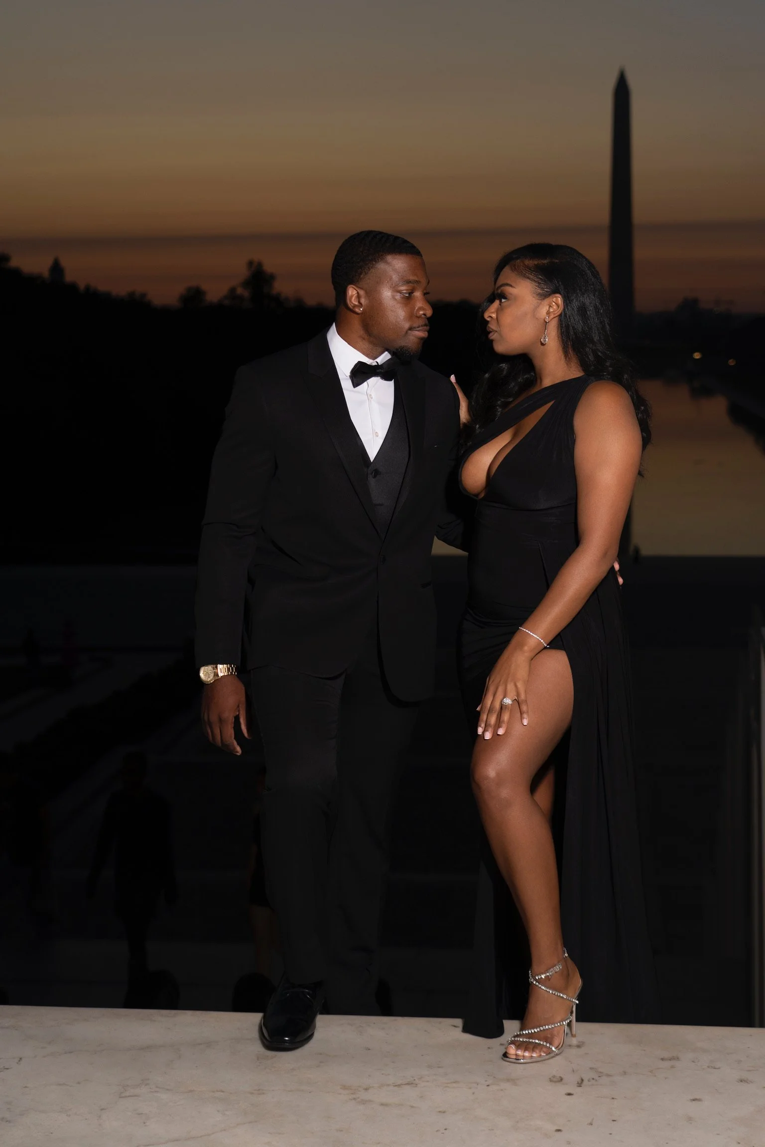 A man dressed in a black tuxedo and a woman in a black evening gown with a thigh-high slit standing close together outdoors during sunset.