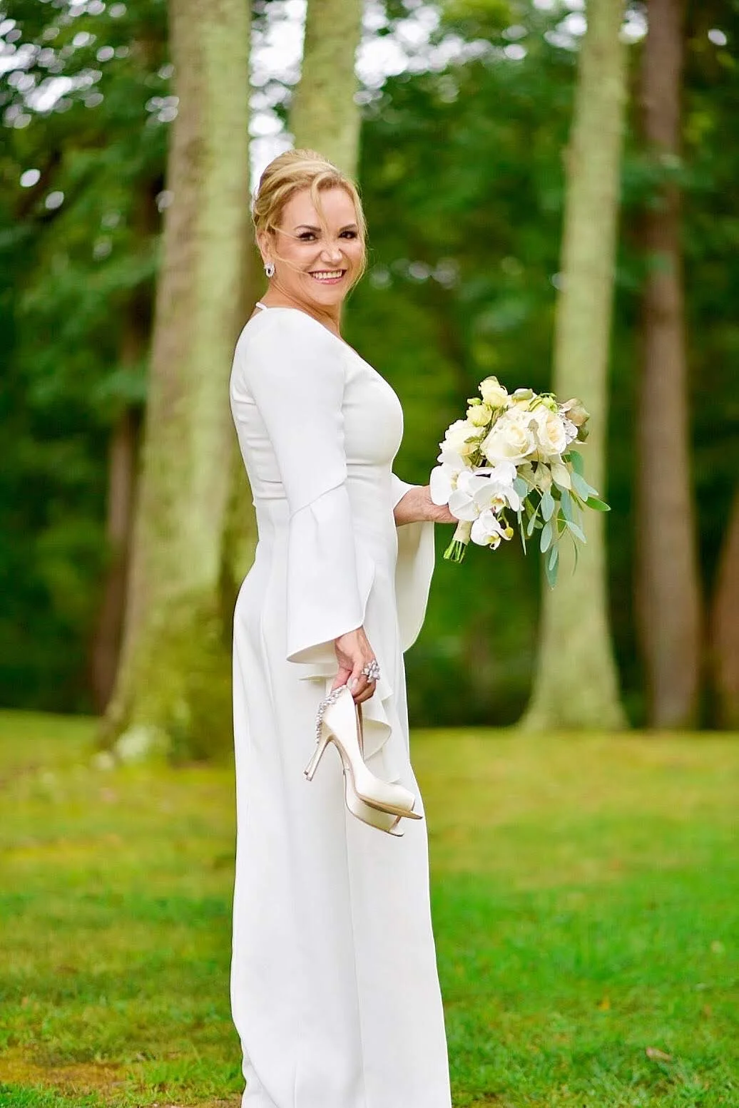 A woman in a white dress holding a bouquet of white flowers and a pair of beige high heels, standing in a lush green forest.