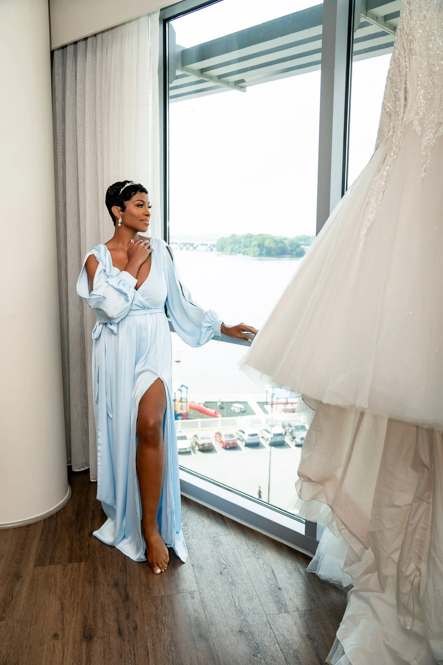 A woman in a light blue gown with a high slit looking at a wedding dress hanging nearby in a bright hotel room with large window overlooking a river and parking lot.