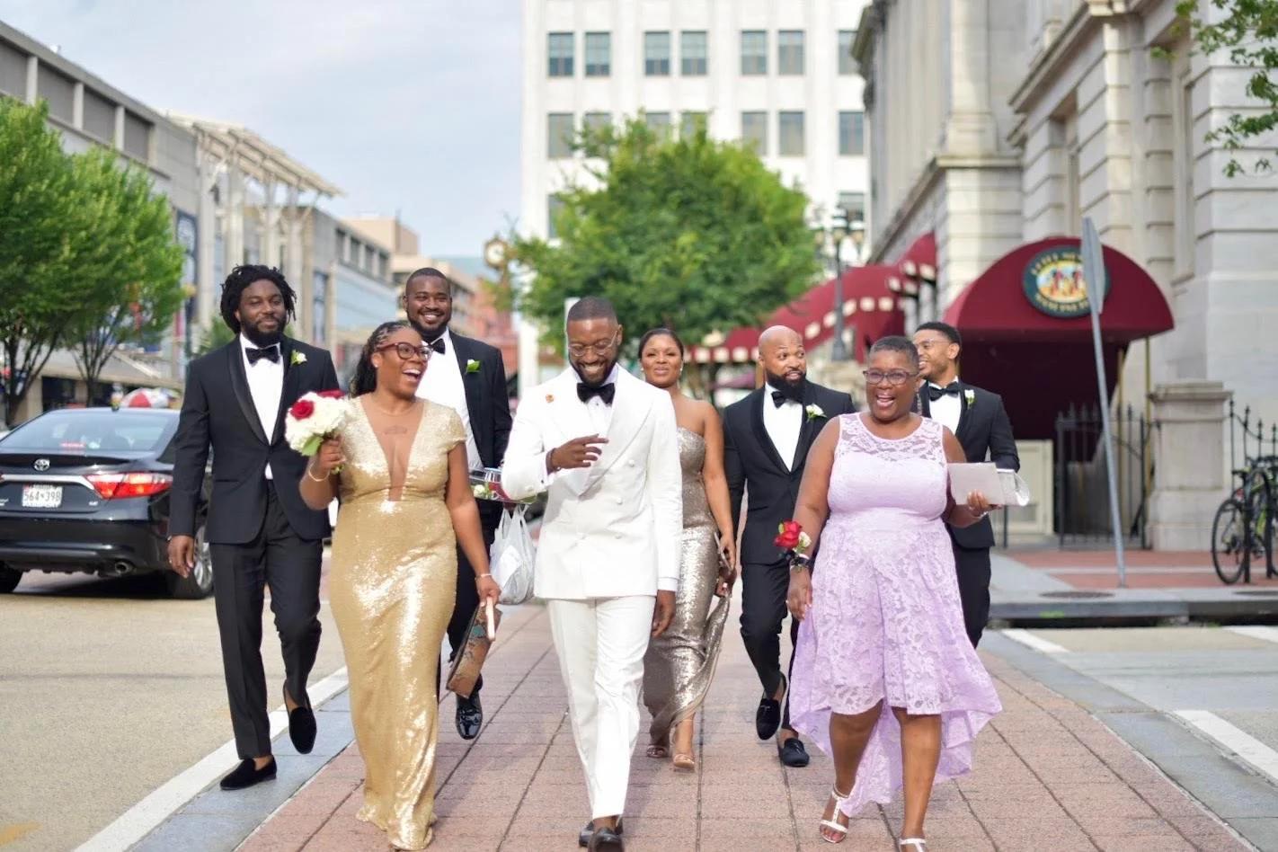 Group of people dressed in formal attire walking on a city sidewalk, celebrating a wedding.