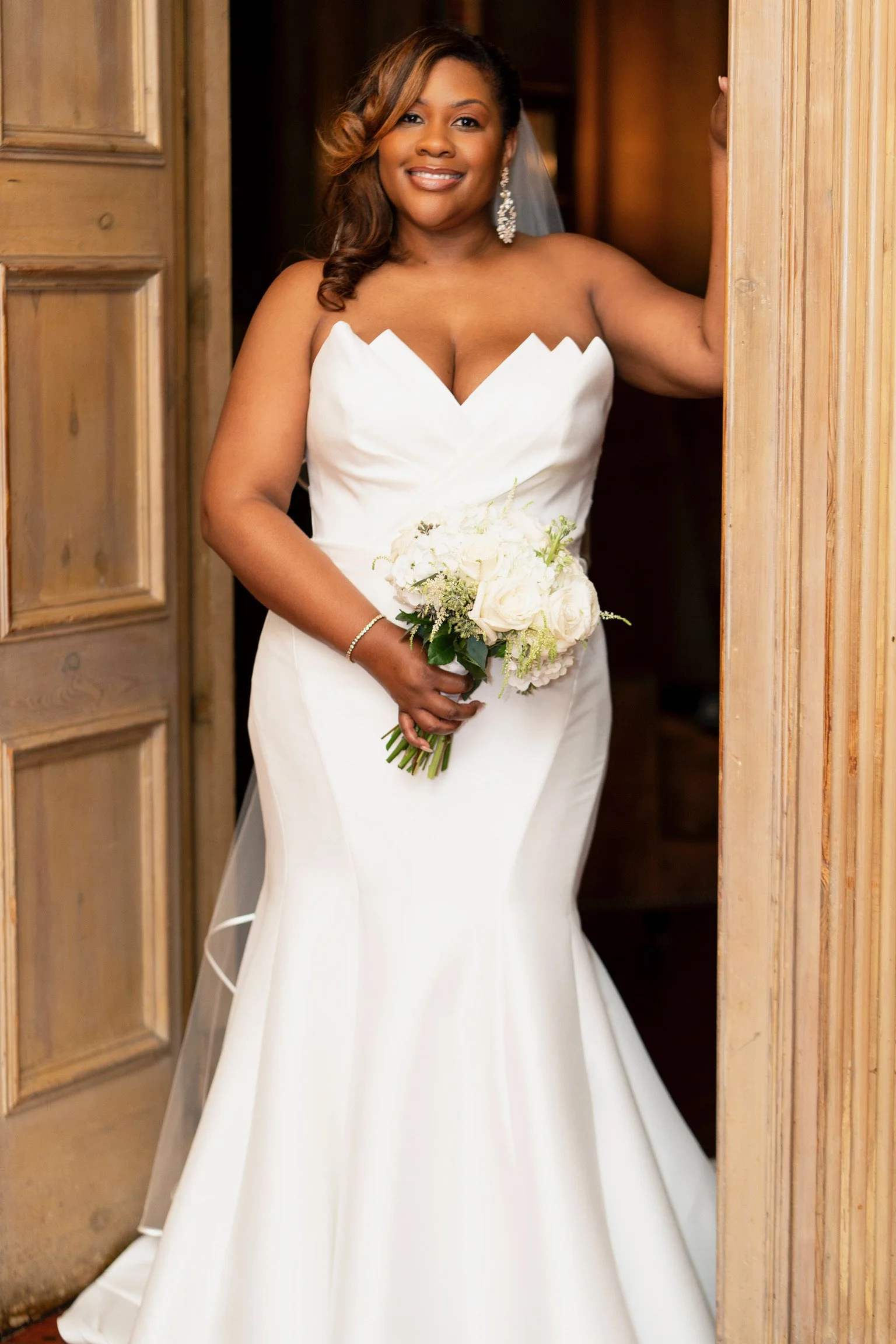 A bride in a white wedding gown holding a bouquet of white roses and greenery, standing in a wooden doorway and smiling.