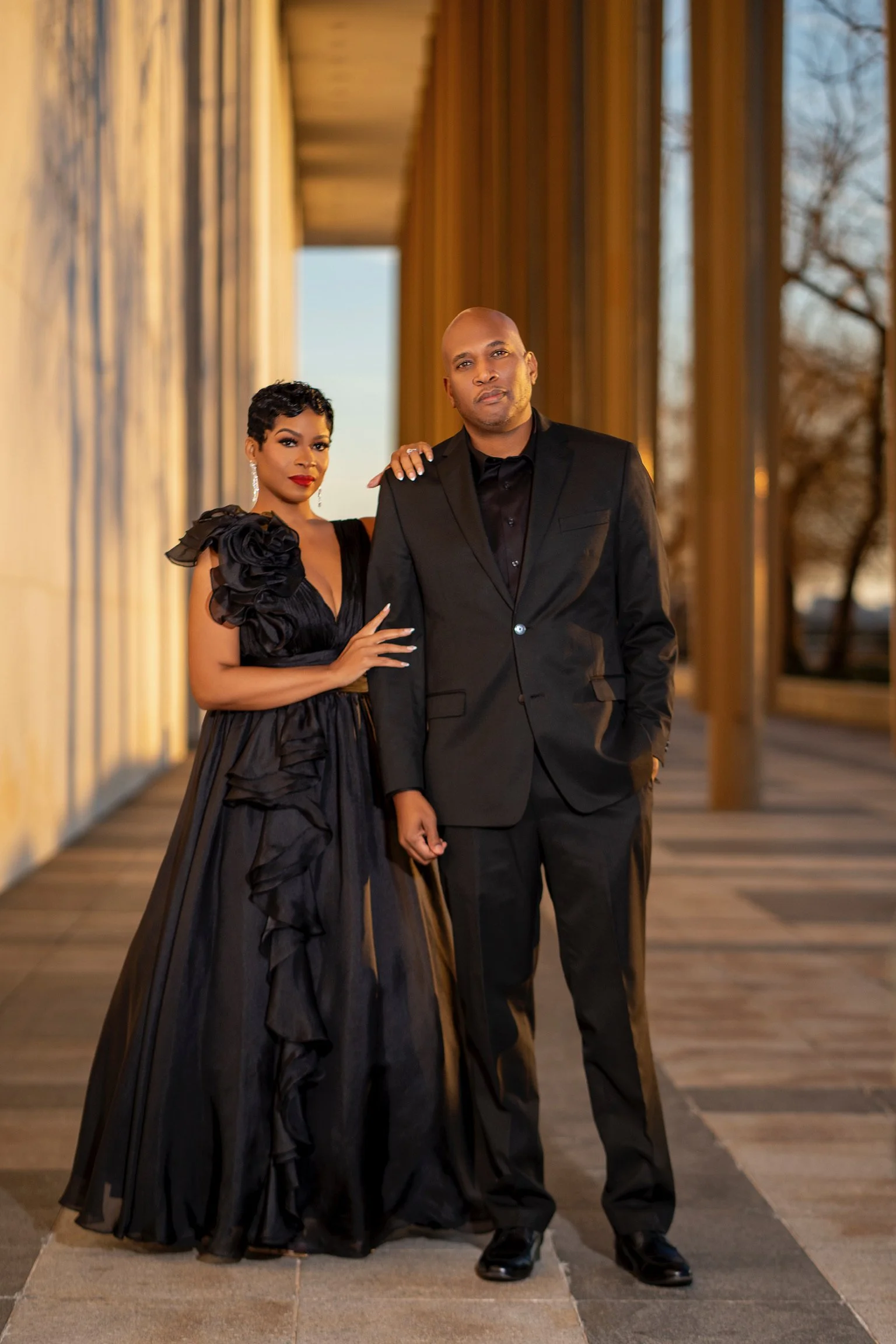A woman in a black gown with shoulder ruffles and earrings stands next to a man in a black suit on a wooden outdoor walkway during sunset.