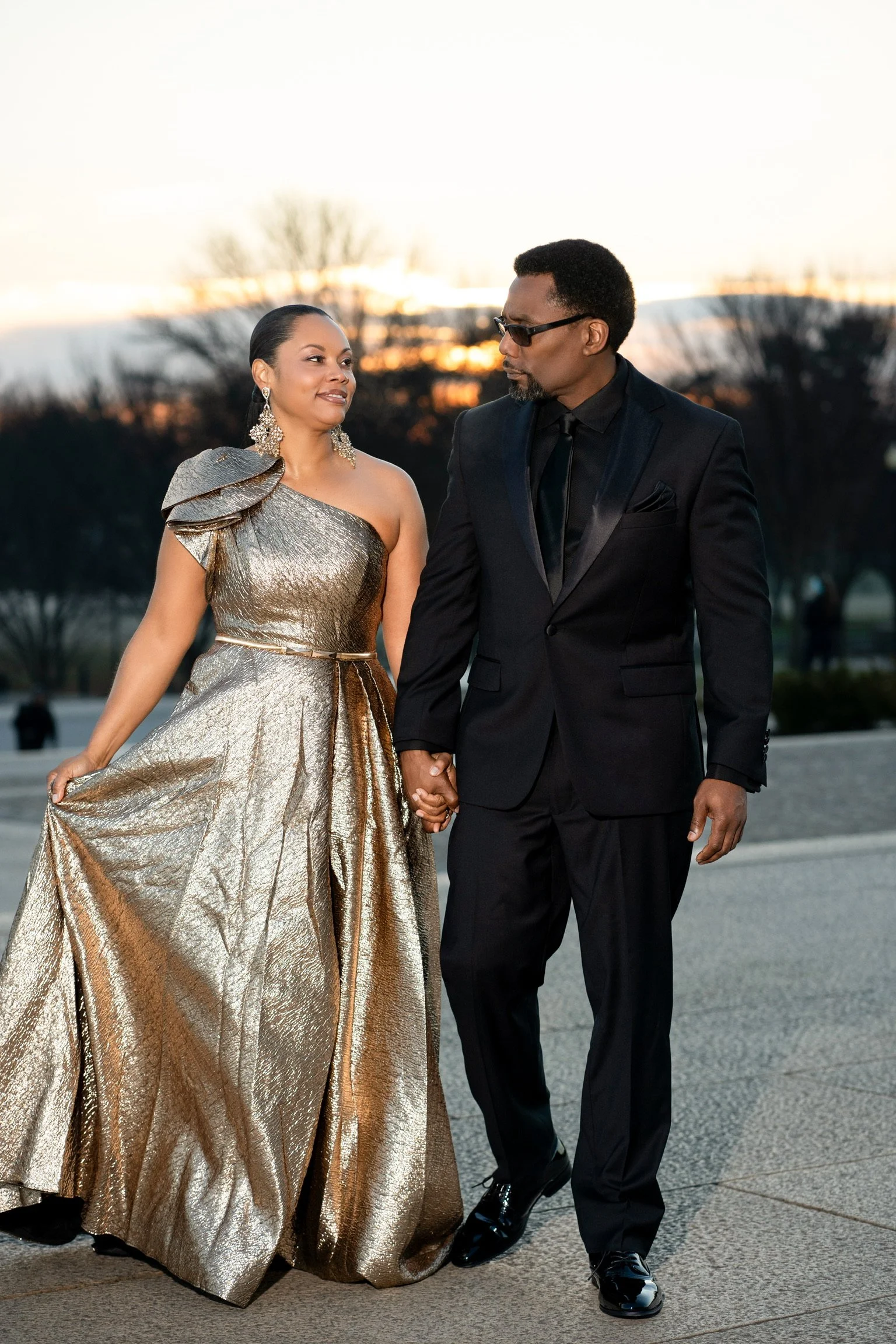 A man and woman holding hands outdoors at sunset. The woman is wearing a gold, one-shoulder, metallic gown with large shoulder detail and large earrings. The man is dressed in a black suit with a tie and wearing glasses.