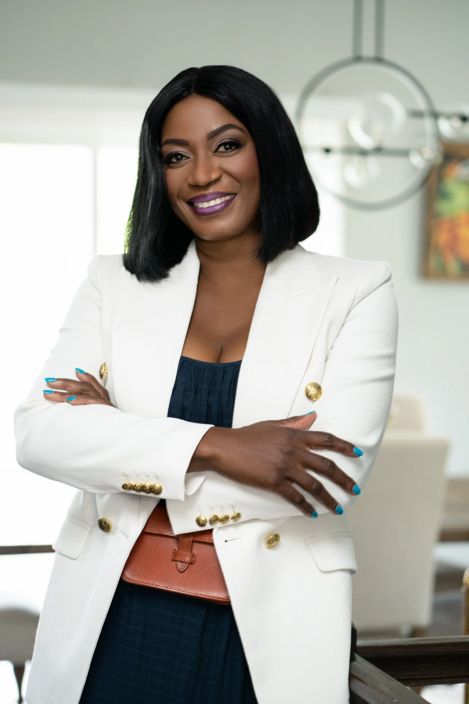 A confident woman with medium dark skin, black hair, purple lipstick, dressed in a white blazer with gold buttons, and a navy top, standing with her arms crossed in an indoor setting with modern decor and artwork.