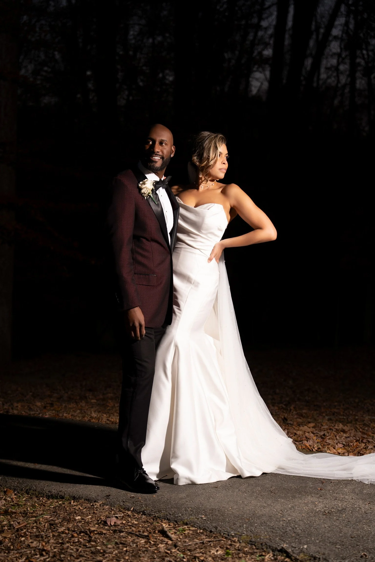 A bride in a white wedding gown and a groom in a suit with a bow tie standing outdoors at night in a wooded area.
