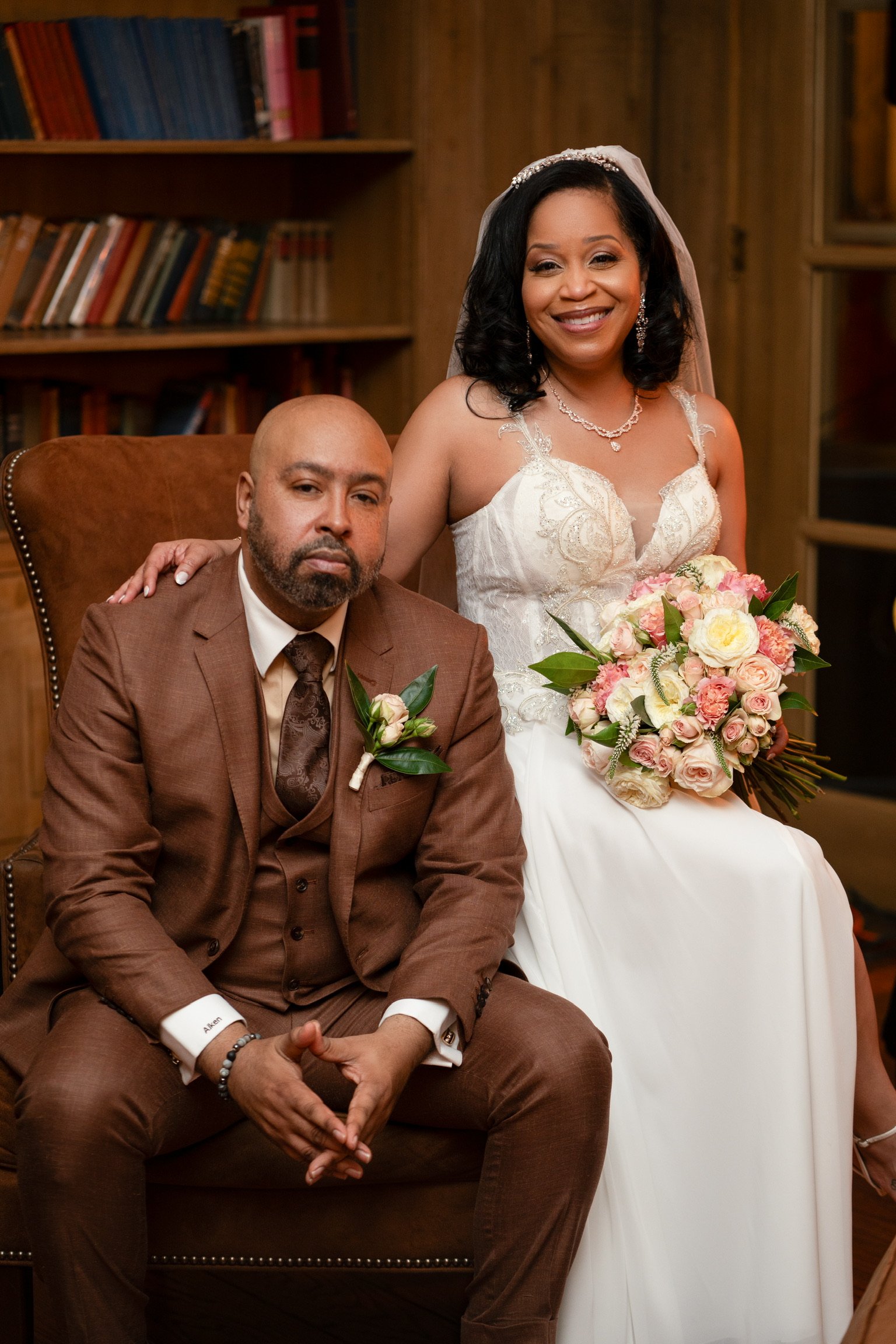 A woman in a white wedding dress and a tiara holding a bouquet, sitting beside a man in a brown suit with a boutonniere, in a wood-paneled room with bookshelves in the background.