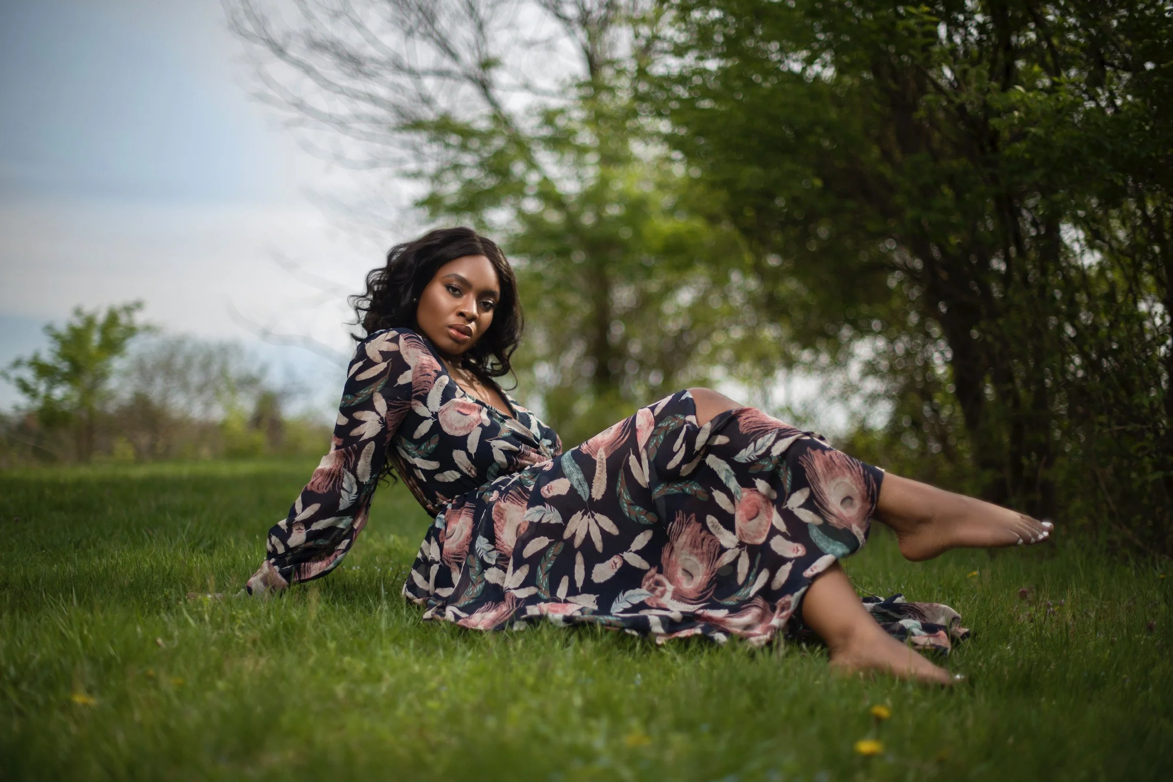 A woman with dark wavy hair relaxing on grass in a park, wearing a long floral dress, with trees and overcast sky in the background.