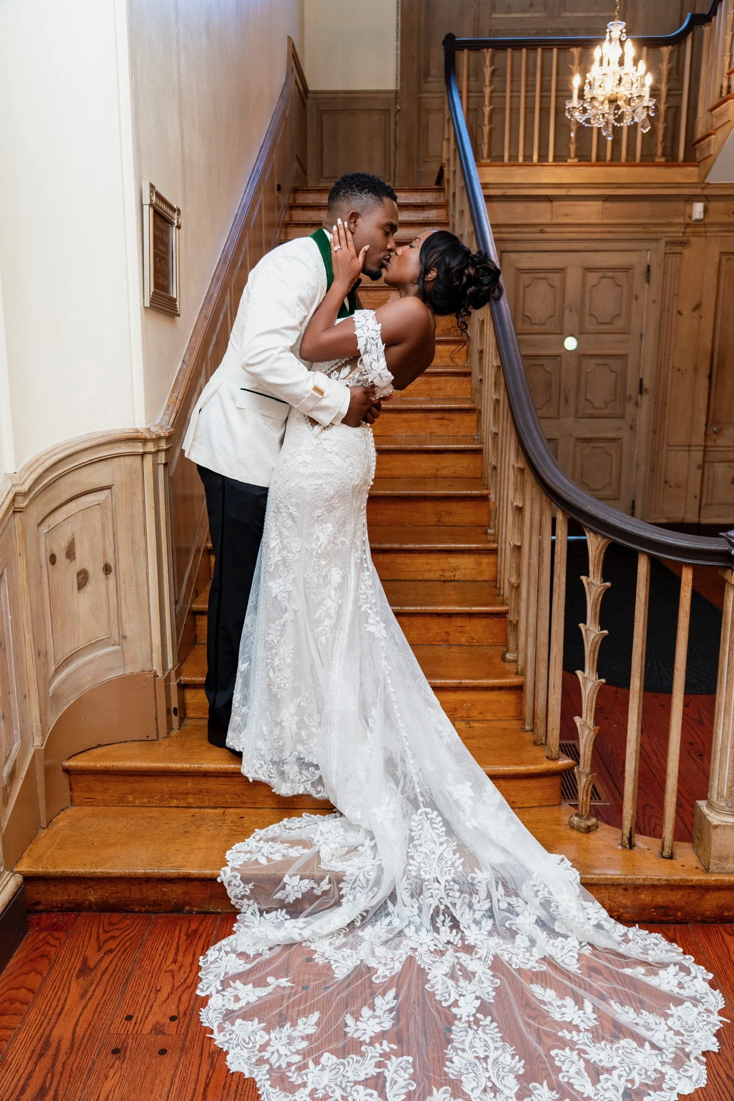 A bride and groom sharing a kiss on a wooden staircase, the bride wearing a lace wedding dress with a long train, and the groom in a white tuxedo jacket with black pants.