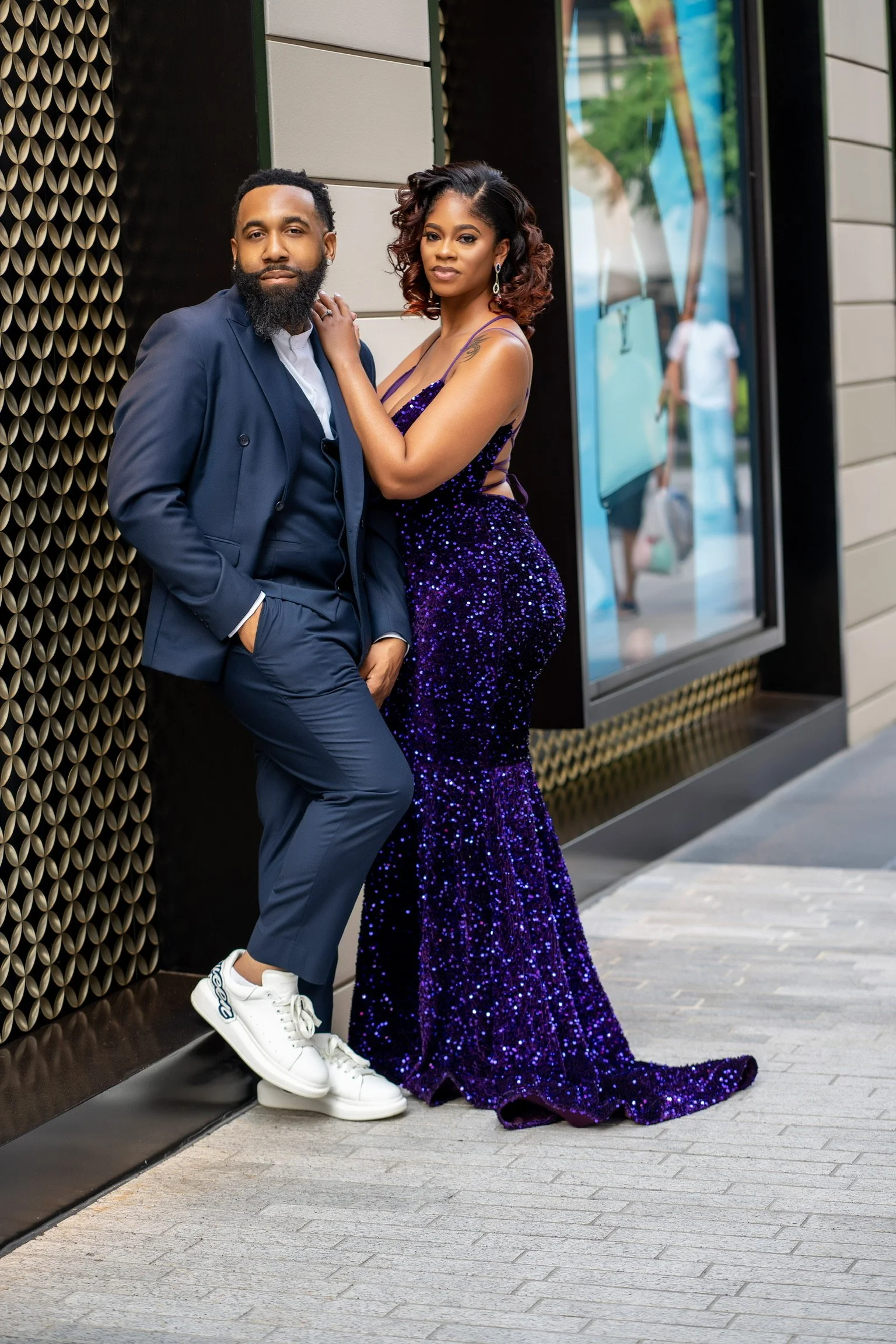 A man and woman dressed in formal attire standing on a sidewalk outside a building with reflective windows, posing for a photo.