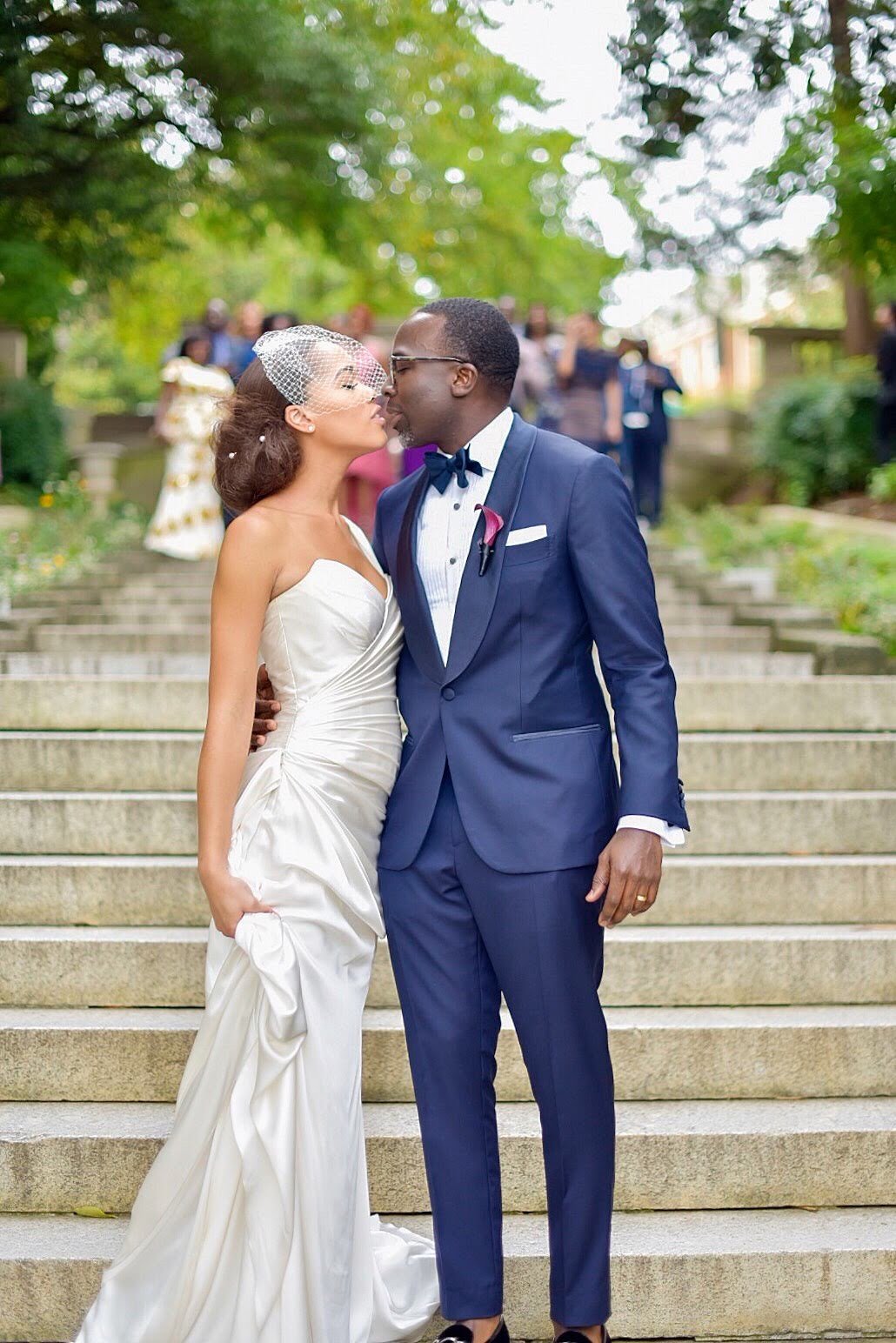 A bride and groom share a moment on the steps during their wedding, with wedding guests in the background, outdoors with trees.