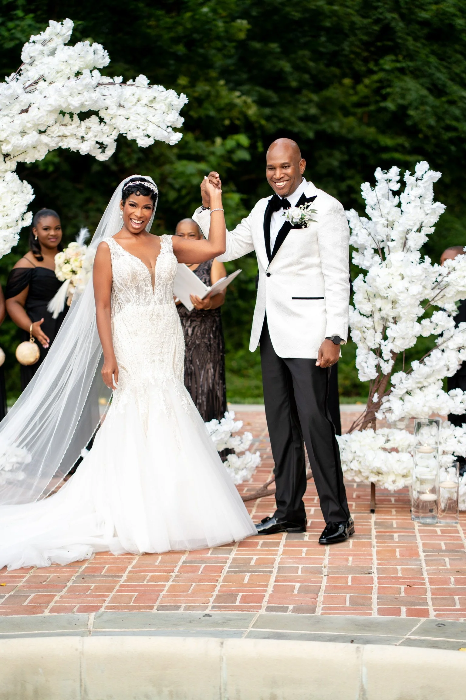 Bride and groom celebrate their wedding outdoors, holding hands. The bride wears a white wedding gown and veil, and the groom wears a white tuxedo jacket with black pants and bow tie. Bridal party members and greenery are in the background.