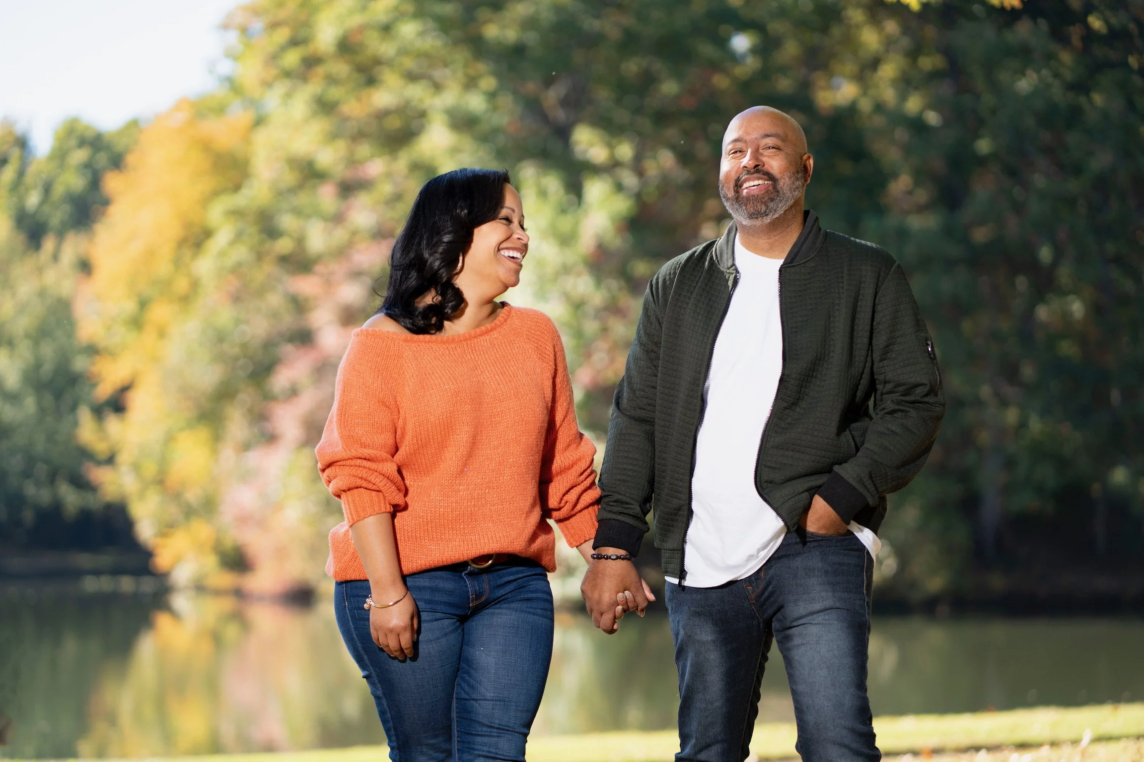 A smiling couple holding hands outdoors near a lake with colorful fall trees in the background.