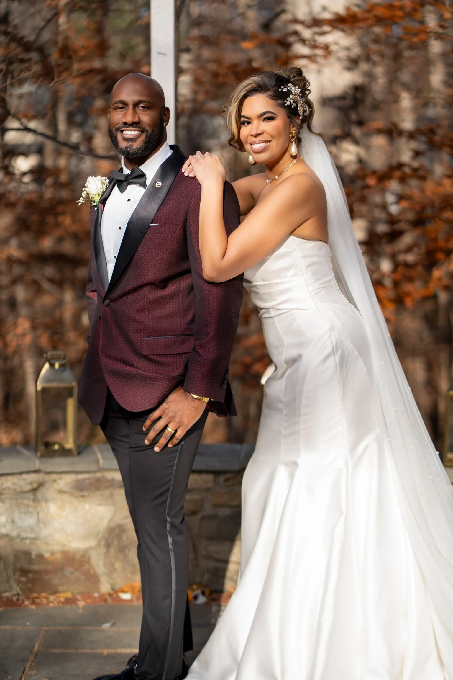 A bride and groom pose outdoors during their wedding, with autumn leaves in the background. The groom wears a maroon tuxedo with black lapels, and the bride wears a strapless white wedding gown with a long veil and floral hair accessory. They are smi