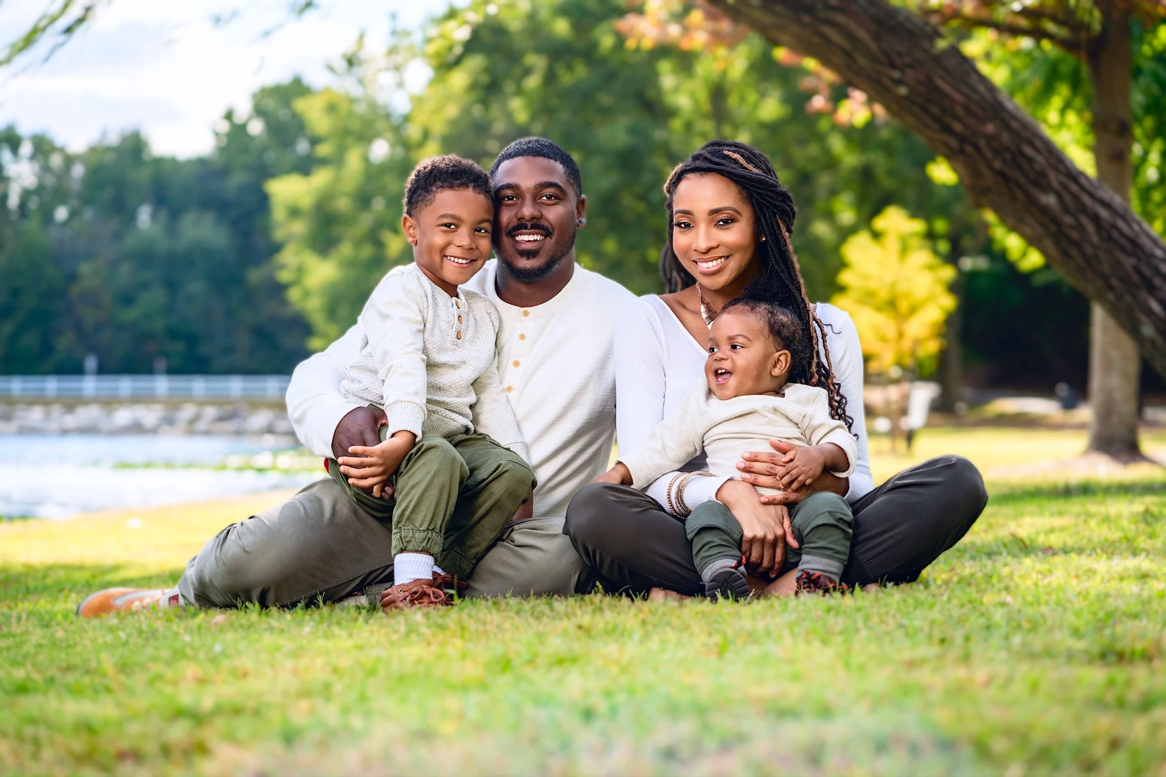 African American family of four sitting on grass in park with trees and river in background, smiling at camera.