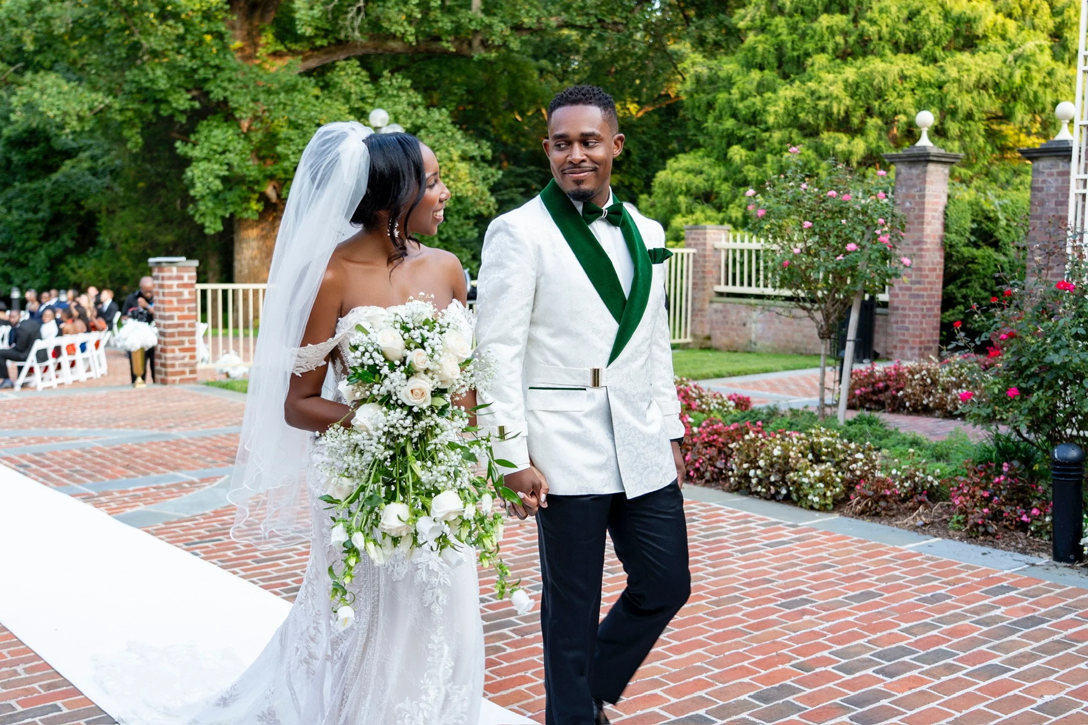 Bride and groom holding hands during outdoor wedding ceremony, with guests seated in the background, surrounded by greenery and flowers.