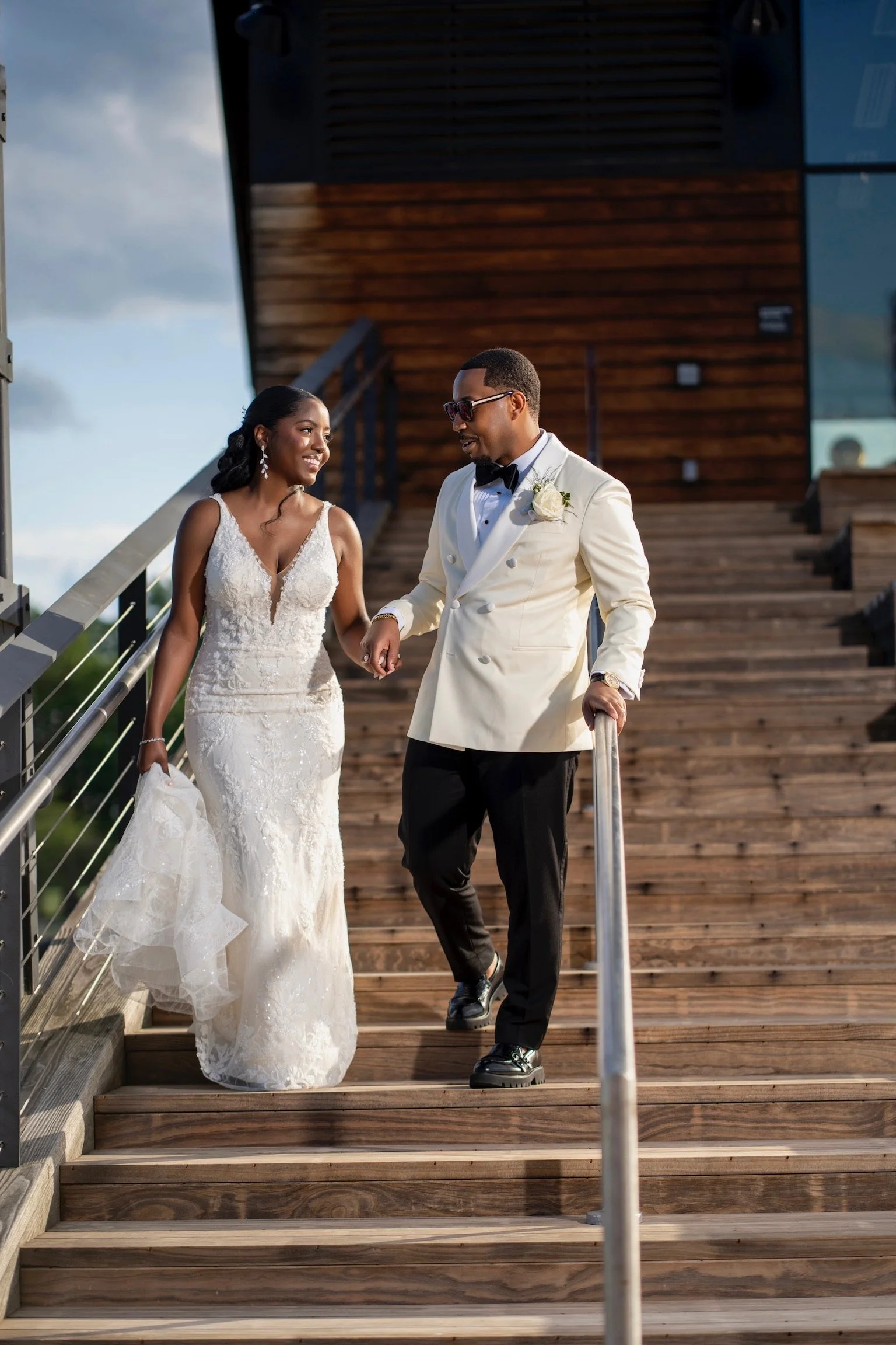 A bride and groom walking down wooden stairs outdoors, holding hands and smiling at each other during their wedding.