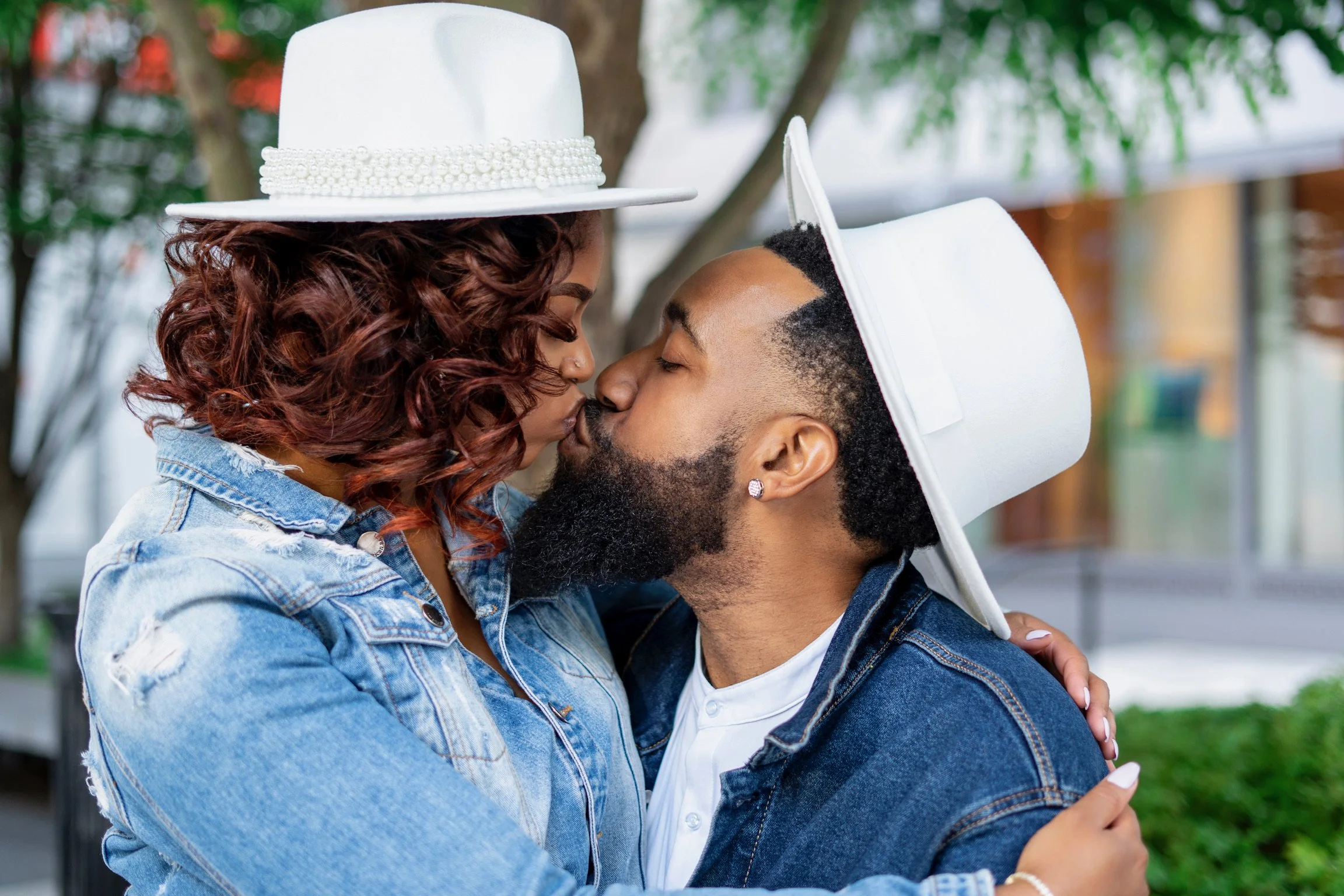 A couple sharing a kiss outdoors, wearing white hats and denim jackets, with trees and buildings in the background.