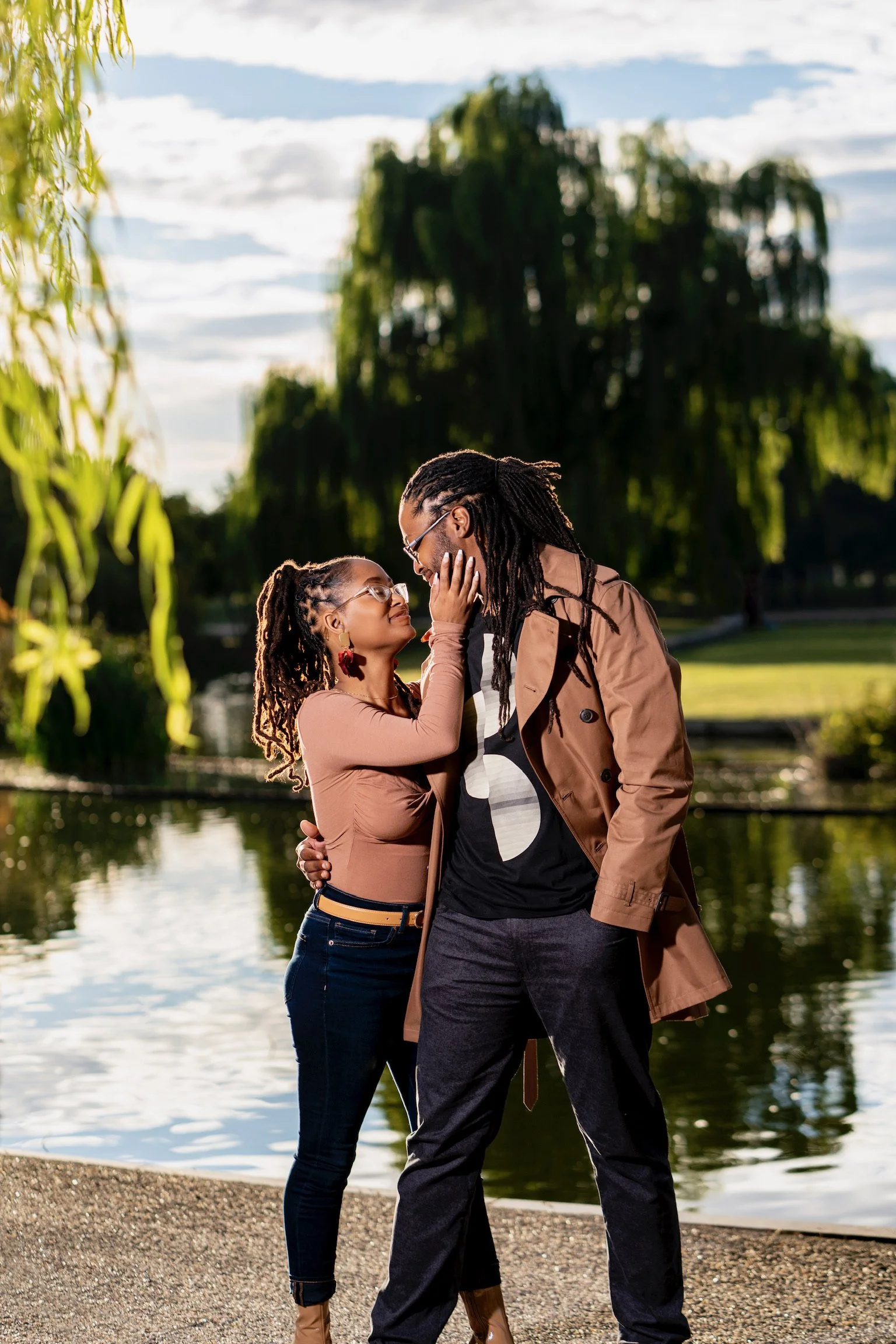 A couple with dreadlocks along with glasses enjoying a romantic moment by a park pond, with trees and sky in the background.