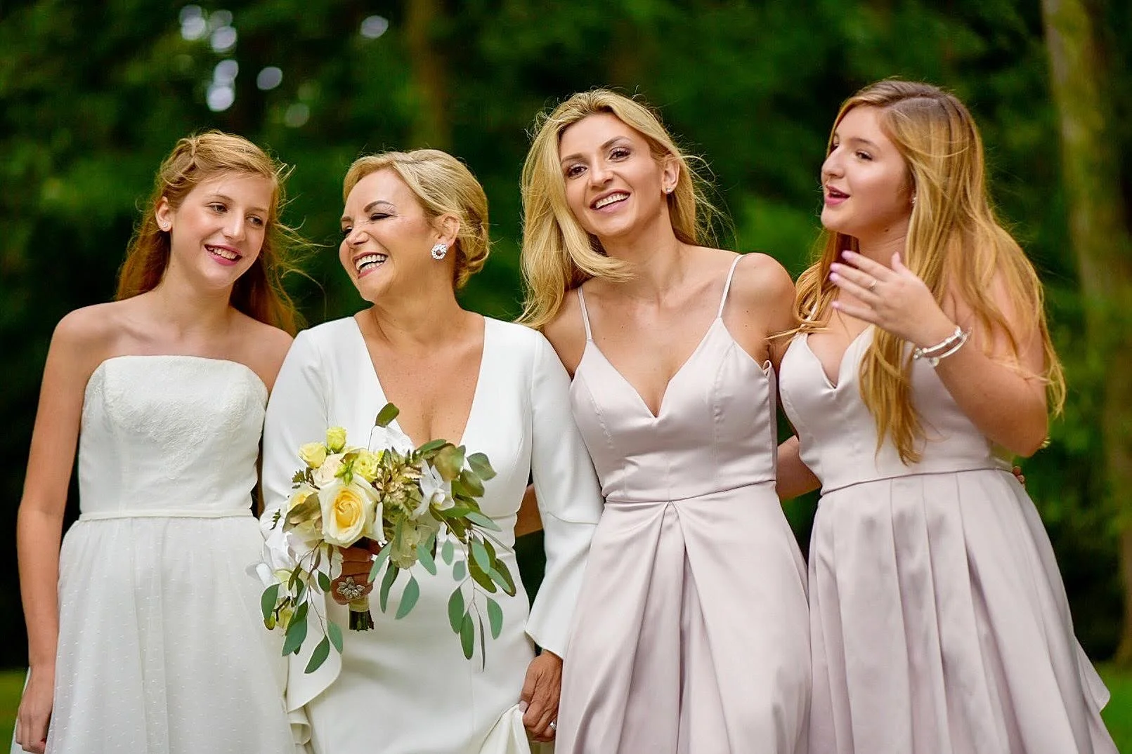 Four women dressed in white and pastel-colored dresses standing outdoors in a green area, smiling and talking to each other.