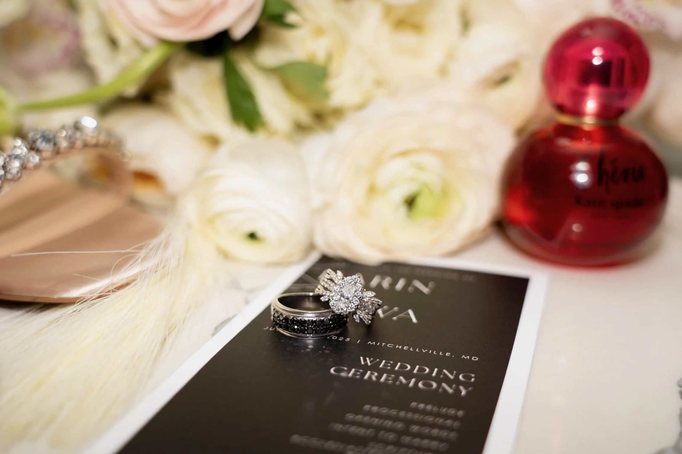 Wedding rings on a wedding program card with white flowers, perfume, and jewelry in the background.