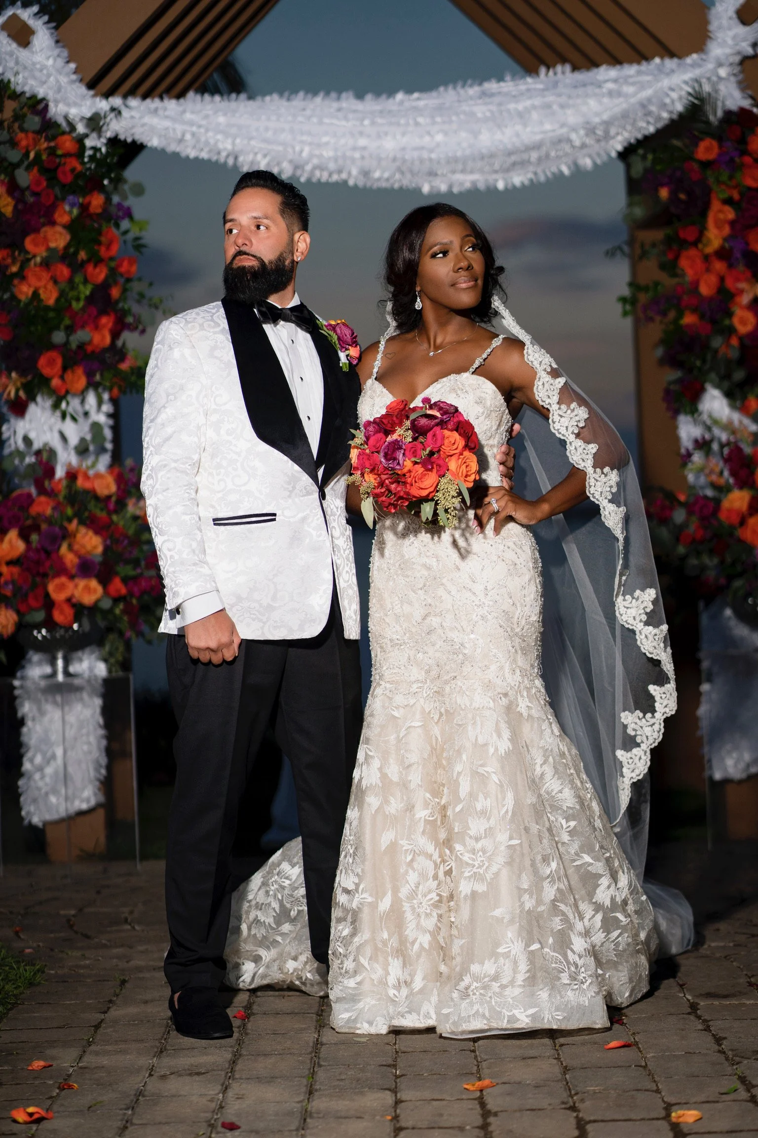 A bride and groom stand together at their wedding ceremony, with the groom wearing a white tuxedo jacket with black lapels and pants, and the bride in a lace wedding gown holding a vibrant bouquet of red, orange, and purple flowers. They are framed b