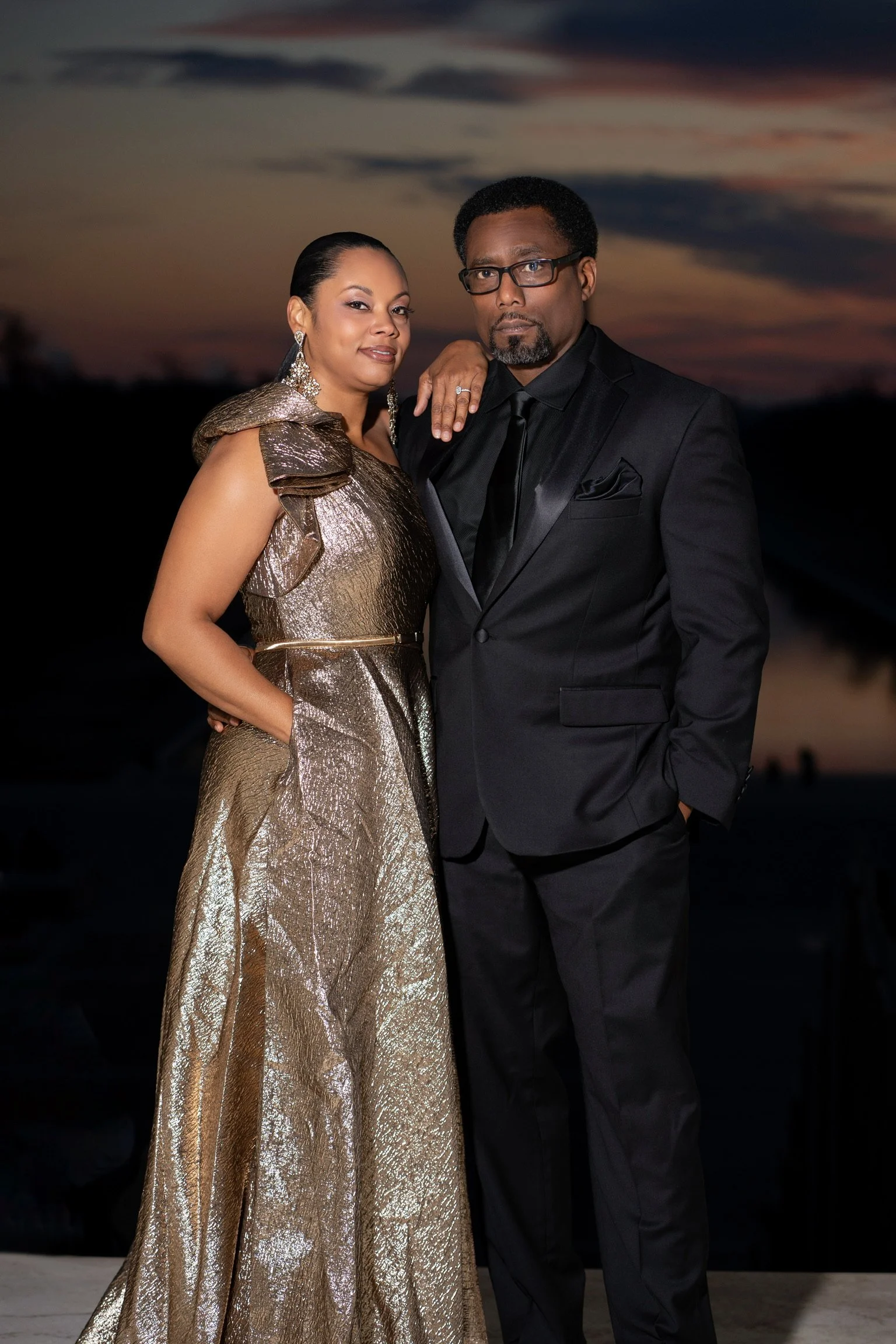 A man and woman in formal attire posing outdoors at sunset.