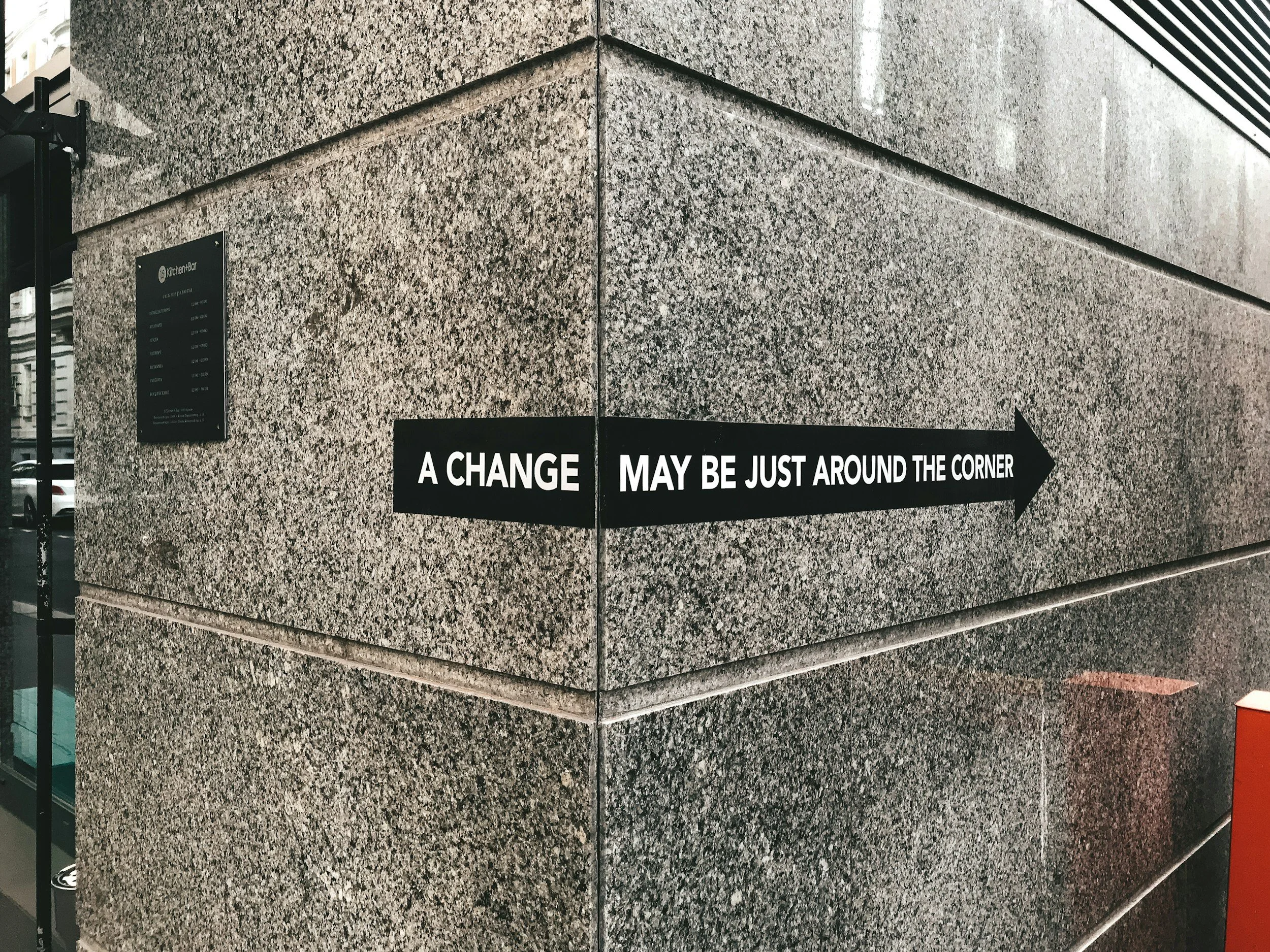 A black arrow-shaped sign on a building corner with white text that reads, 'A CHANGE MAY BE JUST AROUND THE CORNER.'